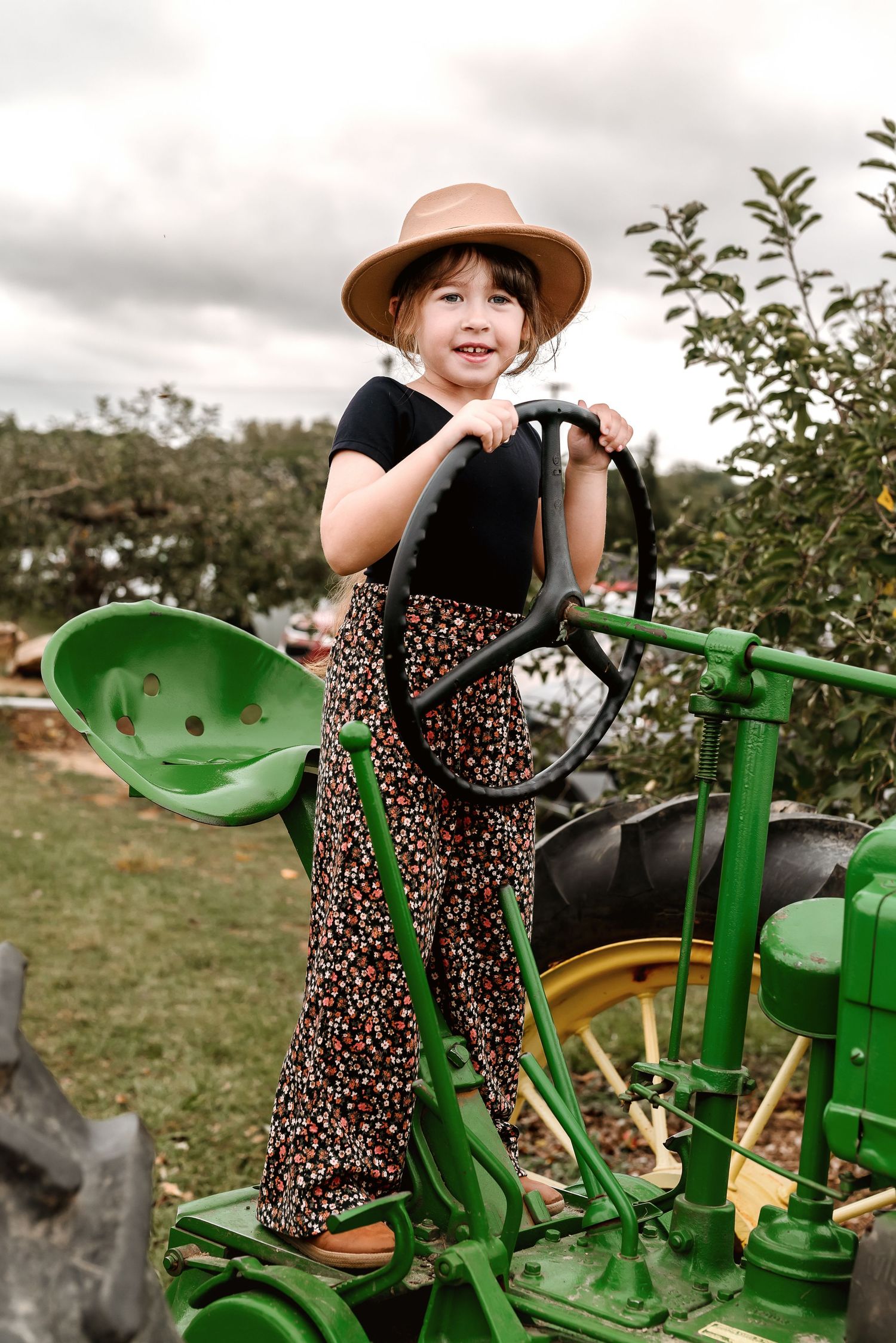 Child in a brown hat and floral dress sits playfully on a vintage green tractor in a sunny field.
