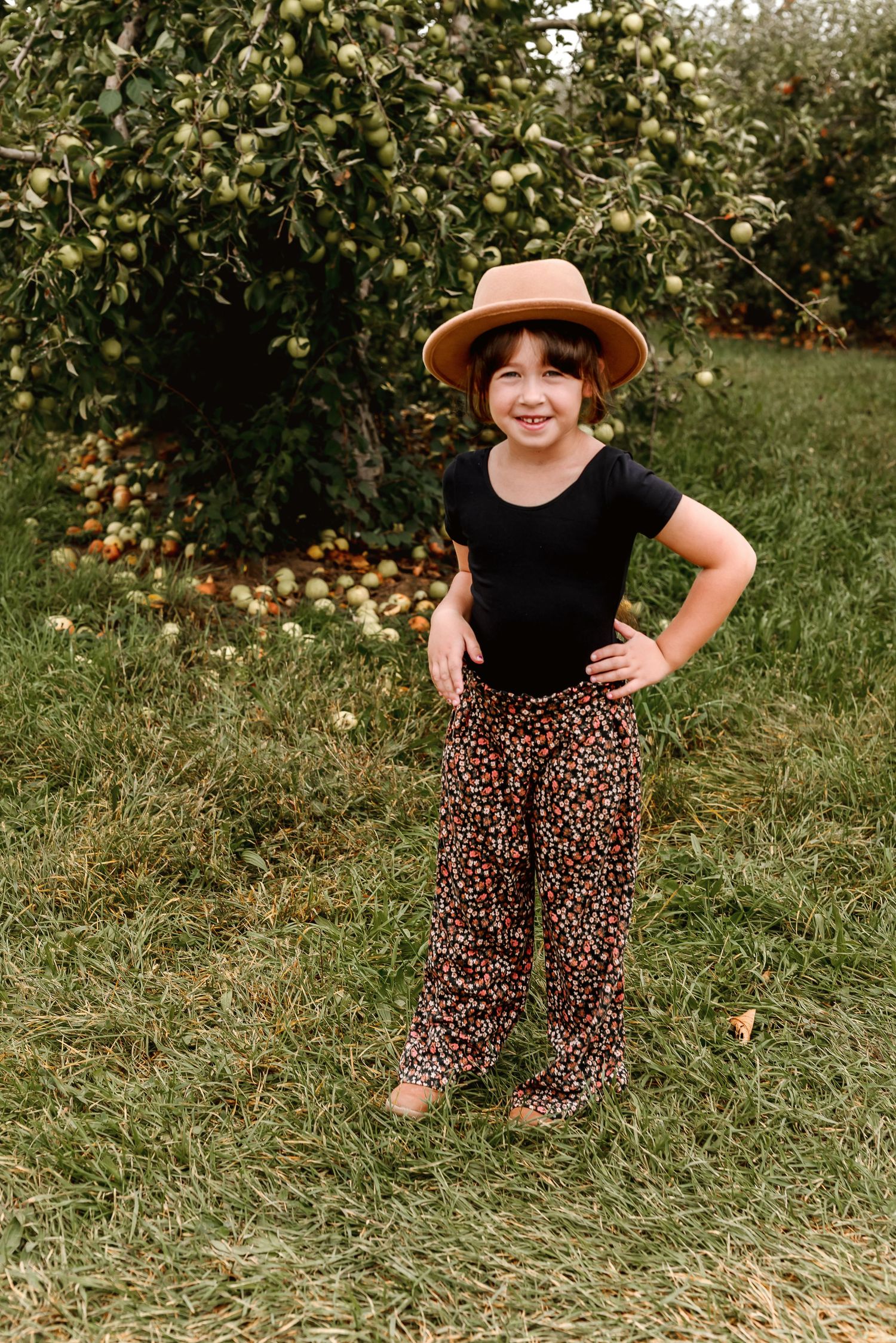 A figure stands in an orange grove wearing a black top, patterned pants, and brown sun hat in a playful pose.
