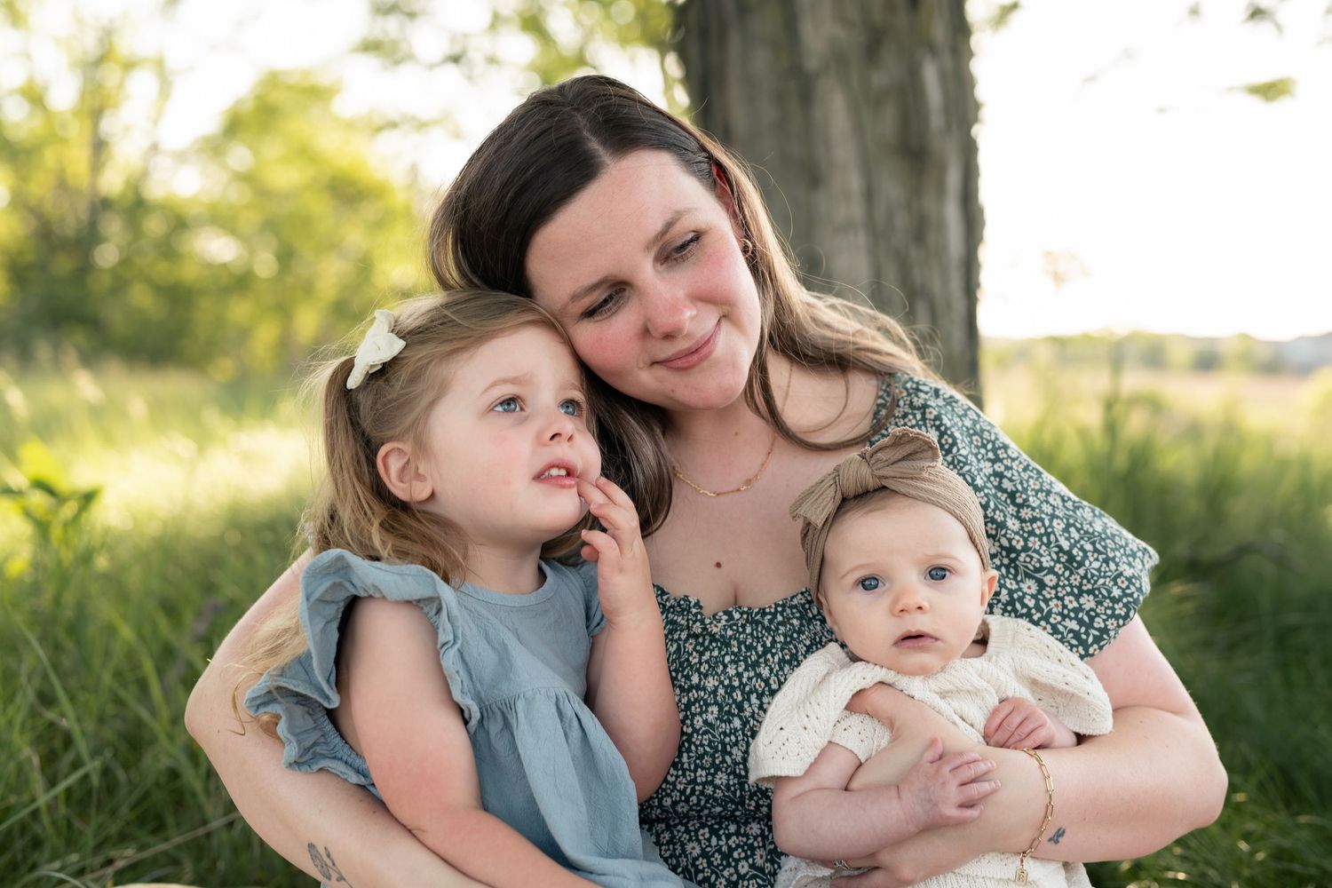 A mother smiles warmly while holding her baby and hugging her young daughter outdoors in a lush green park setting.