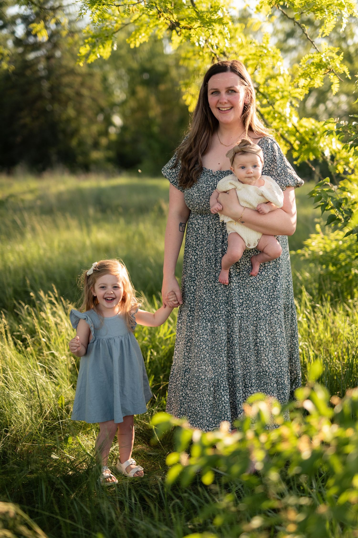 Mother in floral dress holds baby while young girl in blue dress smiles outdoors in sunny green field.