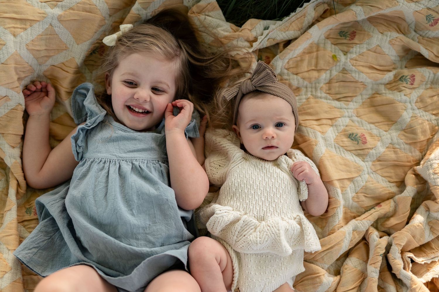 Two young girls lying on a quilted blanket, smiling and wearing cute dresses, captured in a sweet sibling portrait.