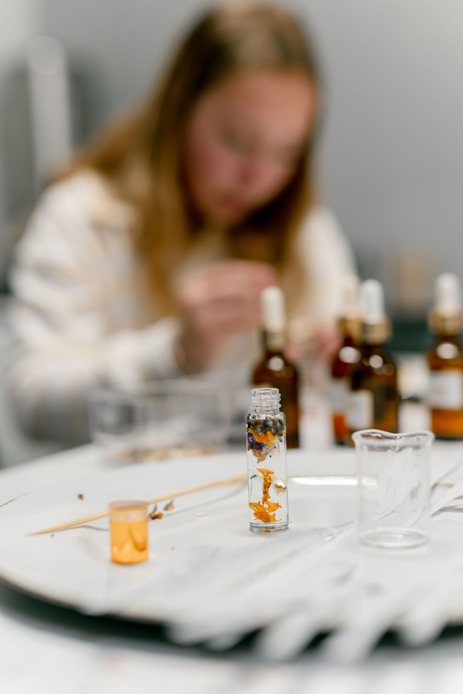 A small glass vial with botanical ingredients on a table, with a blurred woman working on perfume blending in the background.