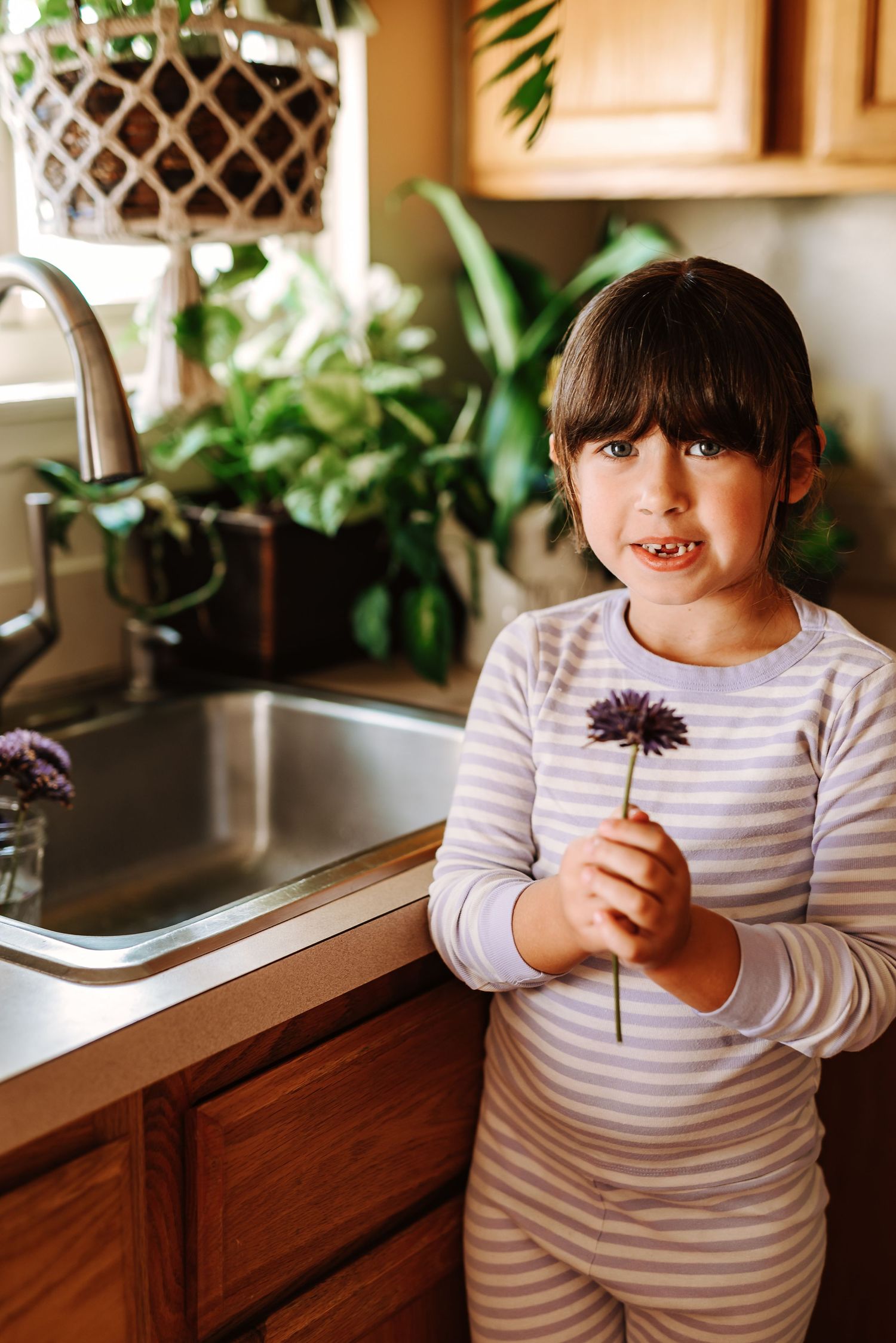 Child in striped shirt holding a purple flower in a kitchen with houseplants and wooden cabinets in background.