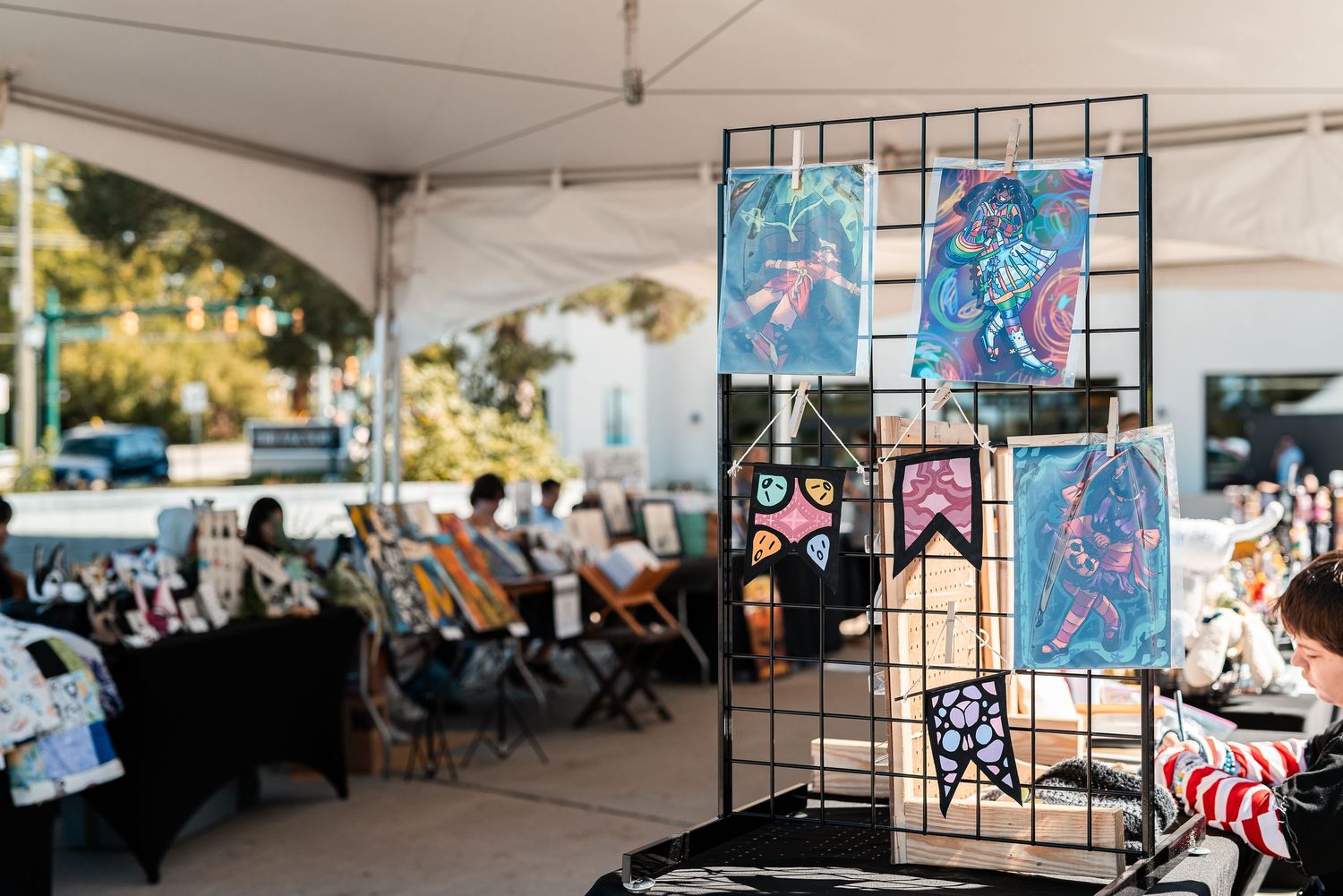 Colorful art prints and handmade items displayed on a grid rack at an outdoor artisan market booth.