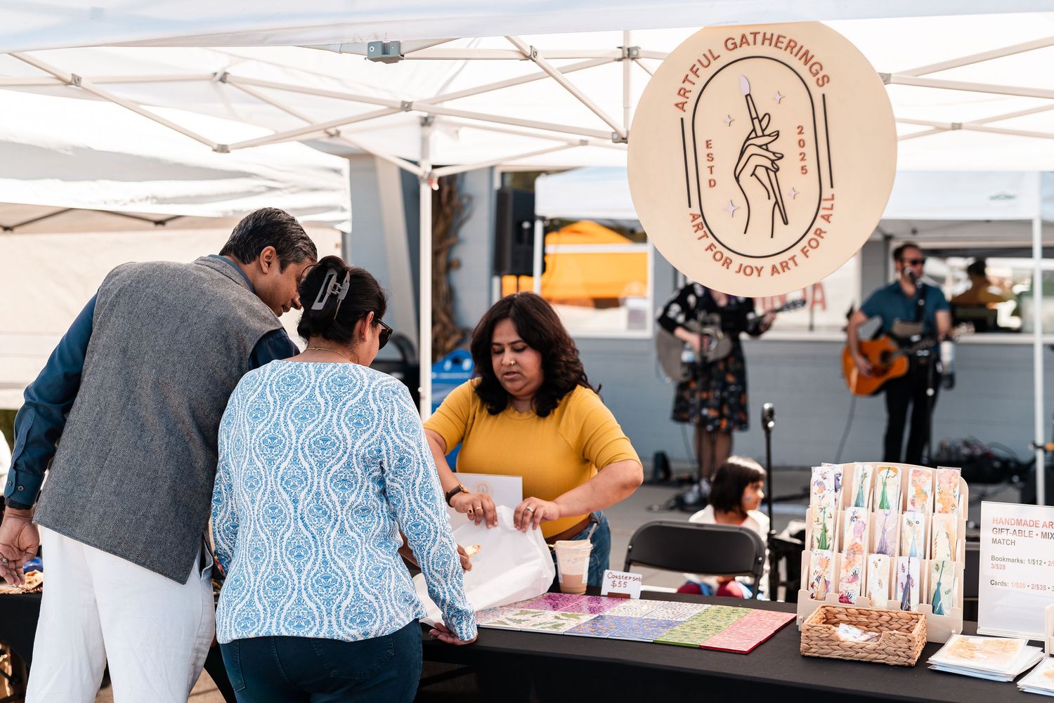 Customers browsing art prints at the Artful Gatherings vendor booth while a musician performs nearby.