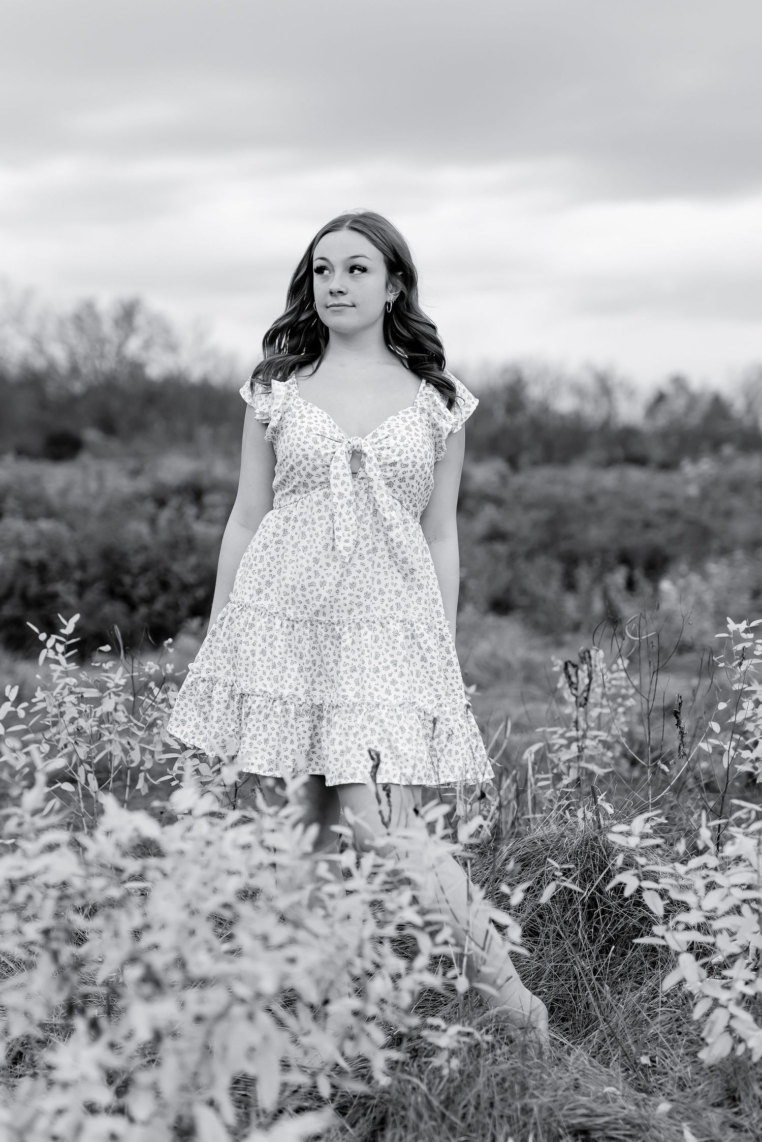 Young woman in floral sundress standing in overgrown field, black and white portrait.