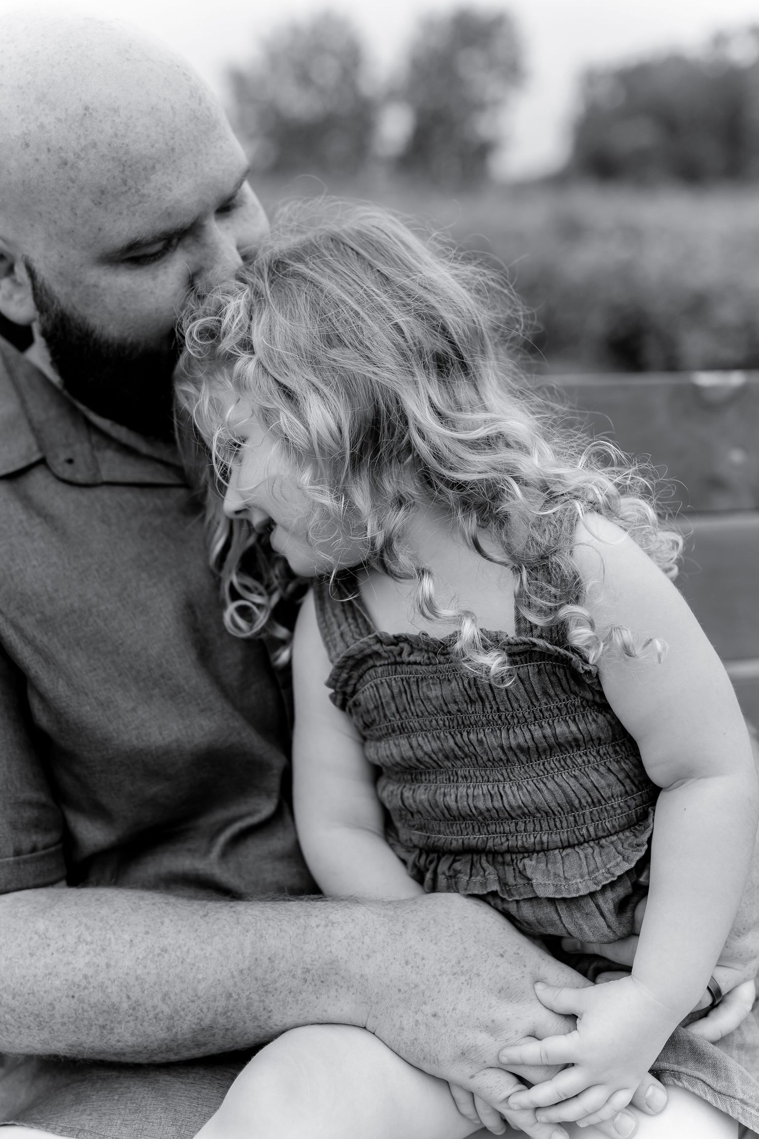 Black and white photo of father kissing curly-haired daughter on a bench outdoors.