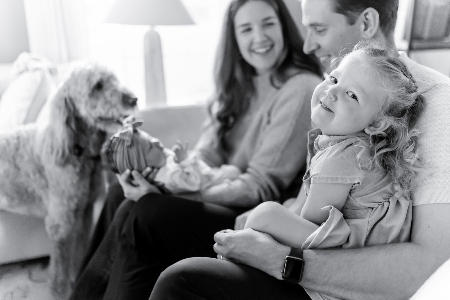 A happy family with a toddler girl, parents, and a dog relaxing together on a sofa at home.