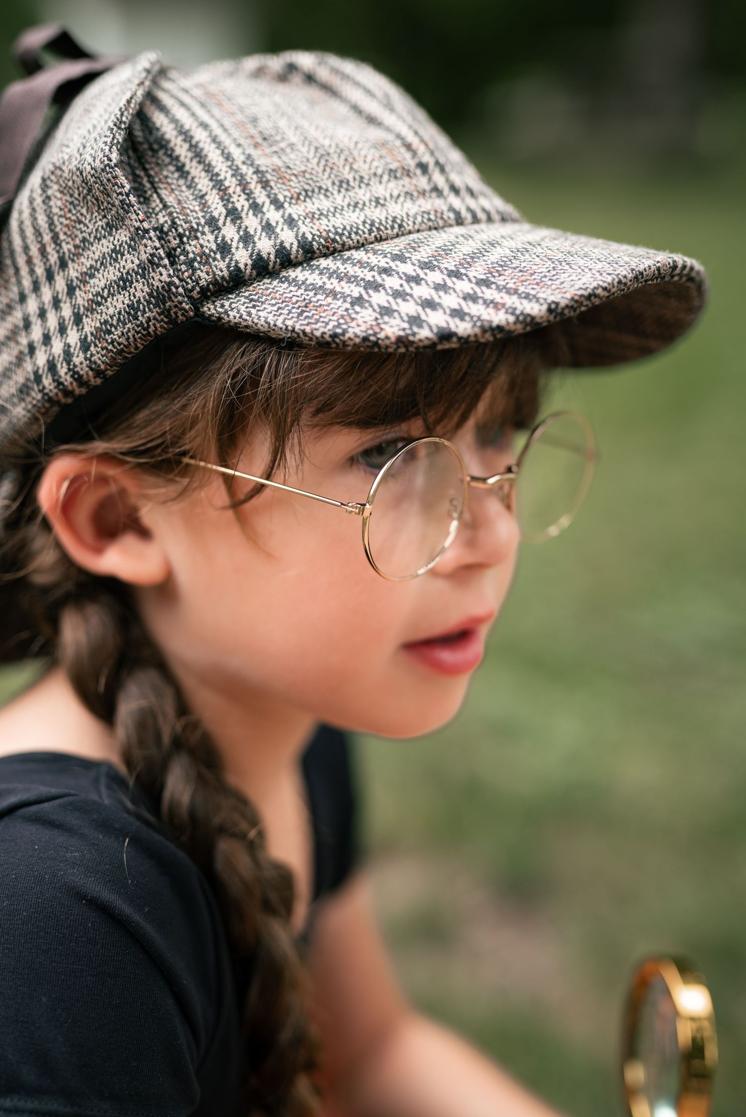 Young girl wearing plaid detective cap, round glasses, and braids holds magnifying glass outdoors.