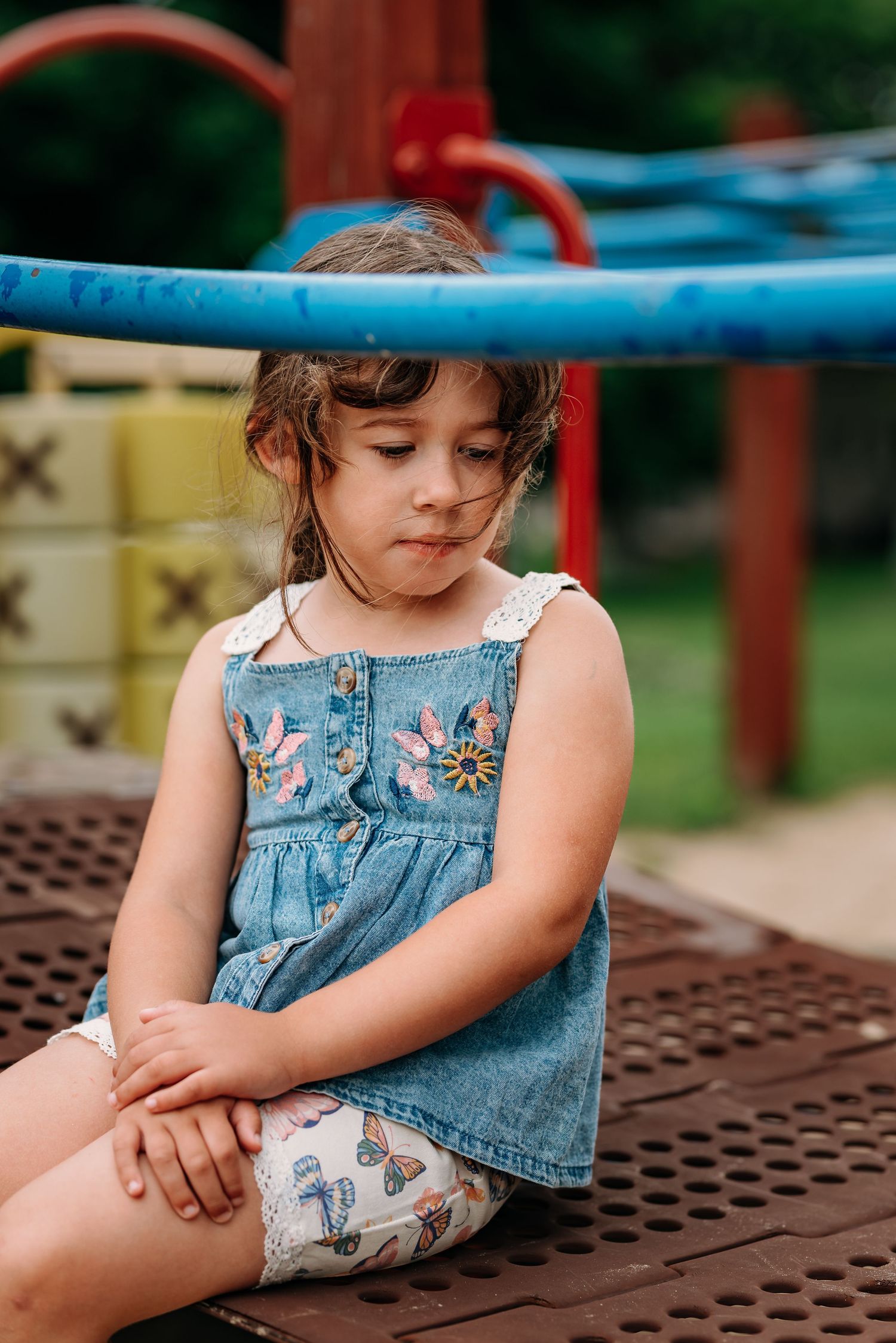 Girl in denim dress leans against a playground structure on a summer day.