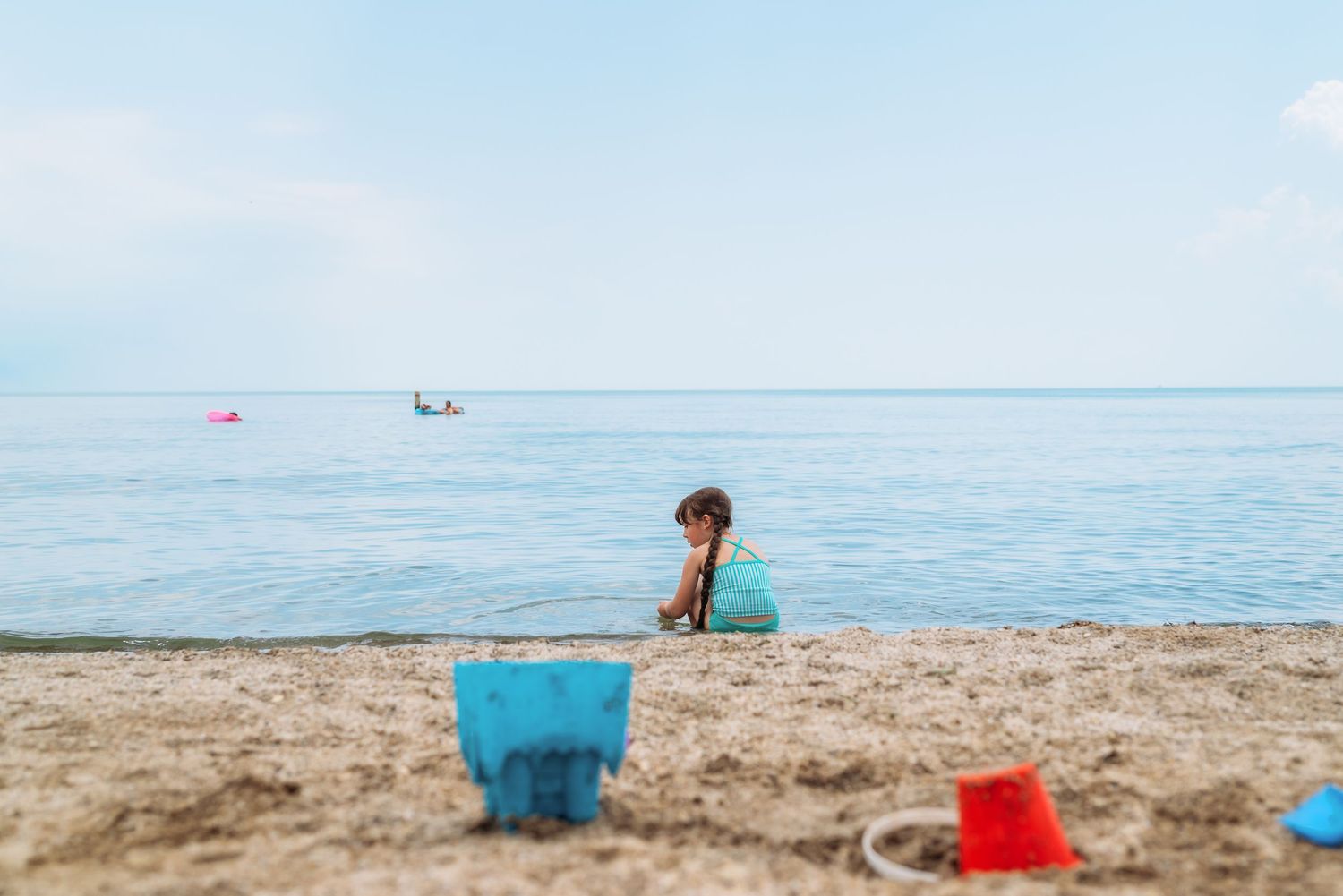 A turquoise bucket and red toys sit on a sandy beach with calm blue ocean waters in the background.