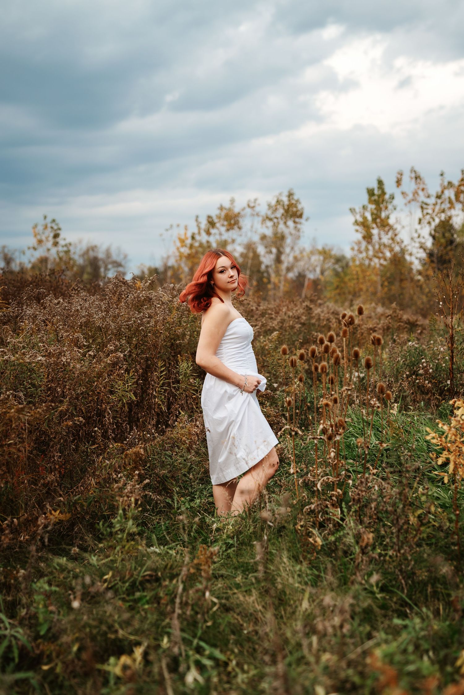 A figure in a flowing white dress twirls through a field of wildflowers and autumn foliage on a cloudy day.