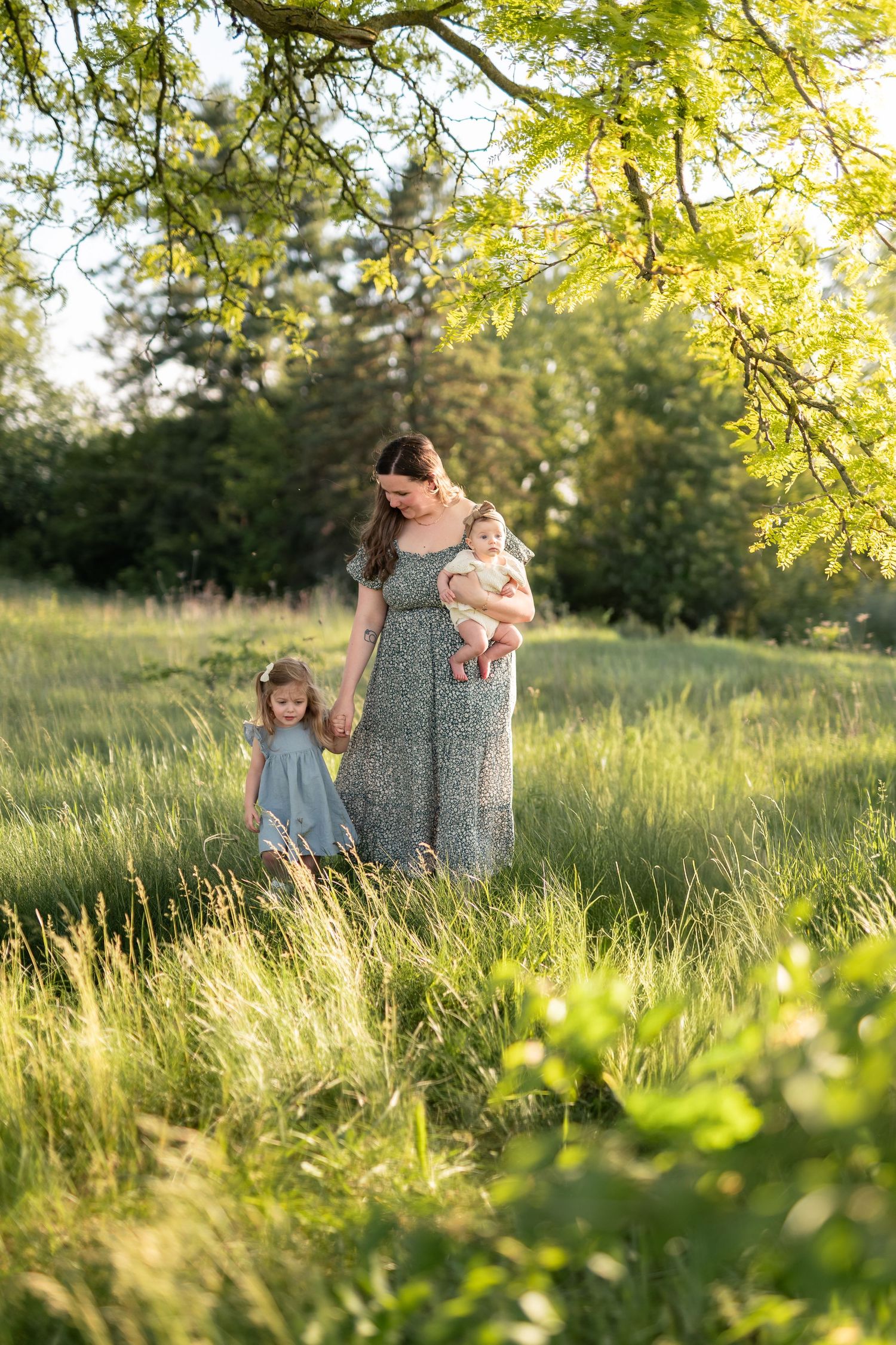 A mother in a floral dress walks with two young daughters through a sunlit green meadow.