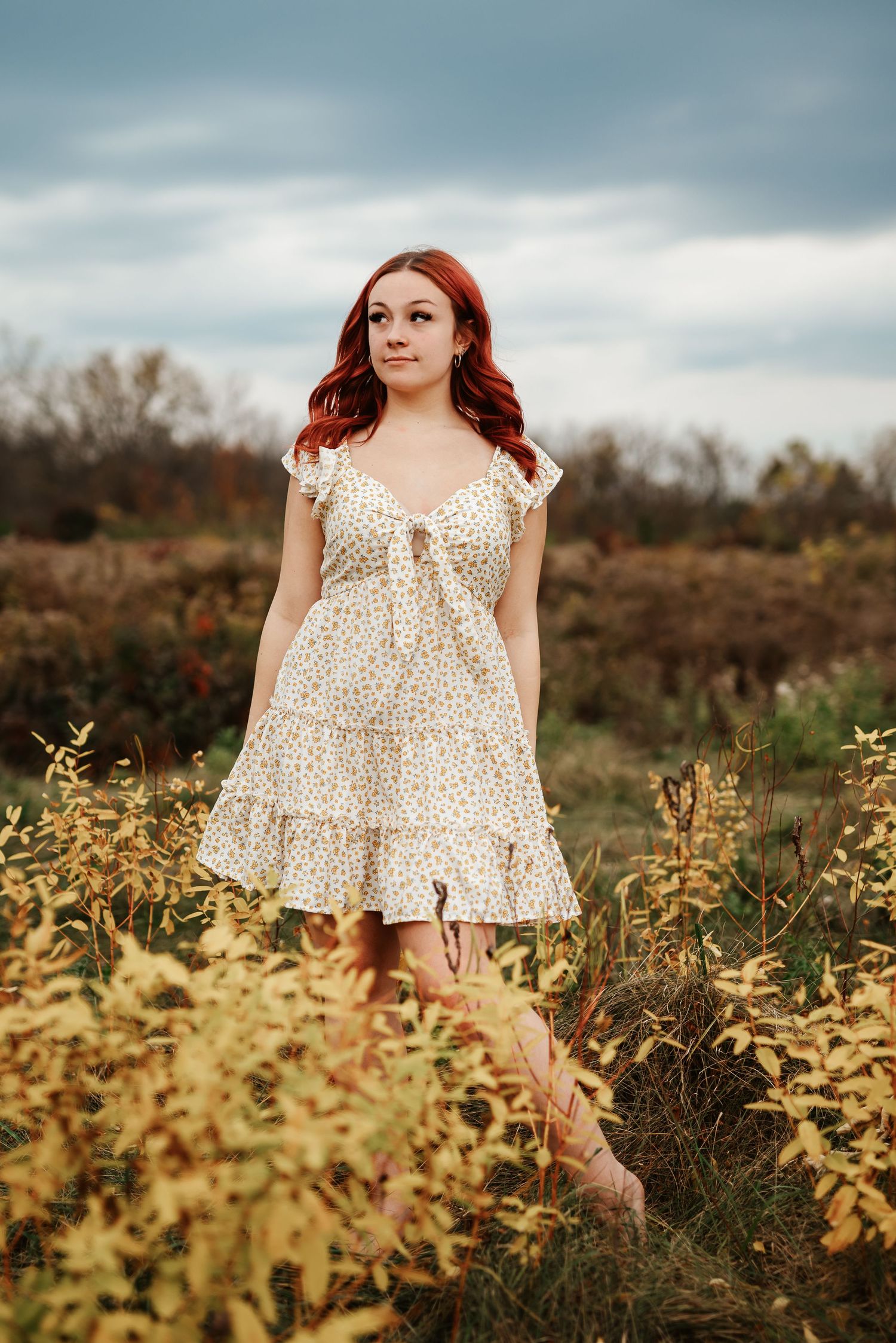 A person in a cream polka dot dress stands in a golden autumn field under an overcast sky.