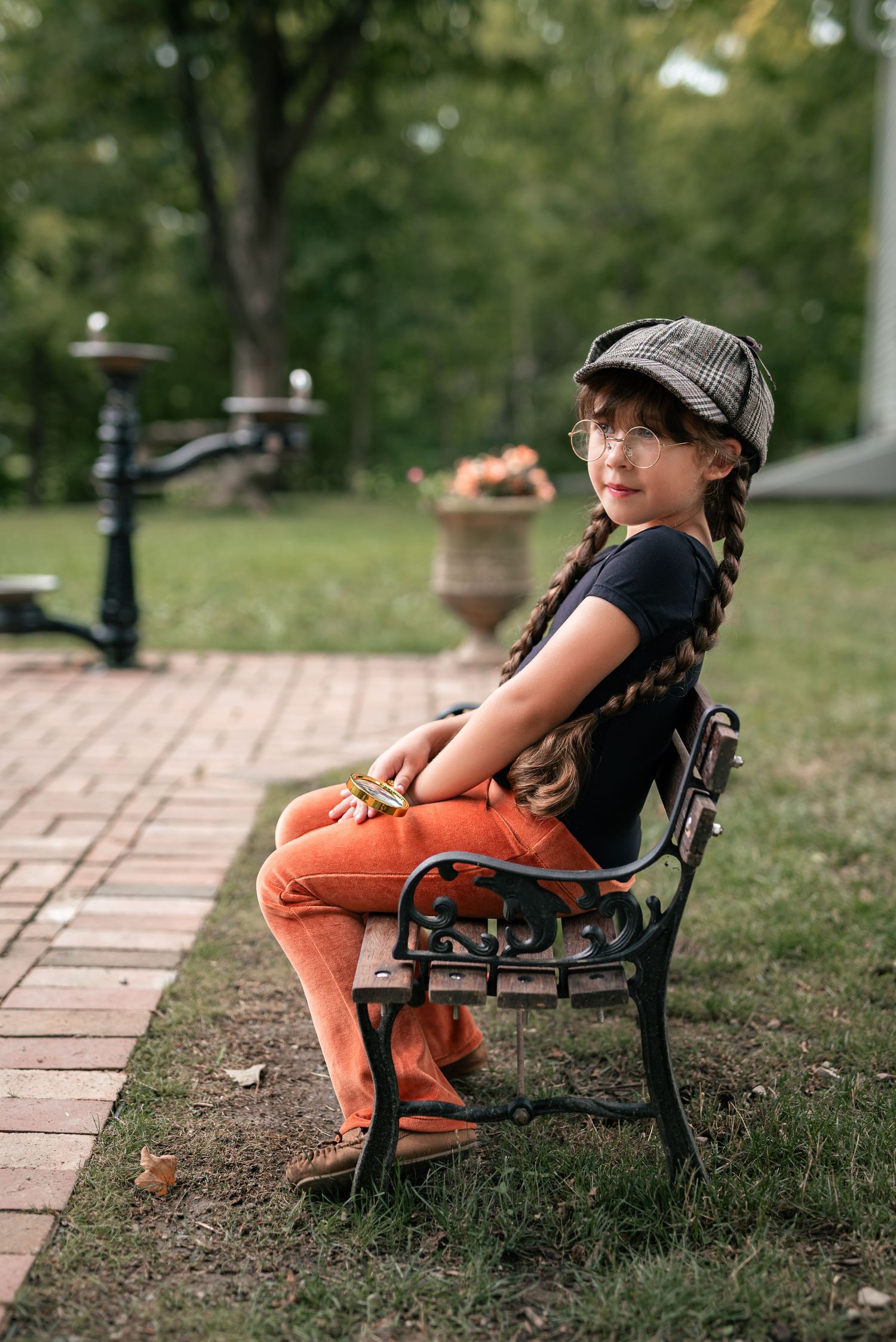 Young girl in orange pants and newsboy cap sits on ornate iron bench in lush green park setting.