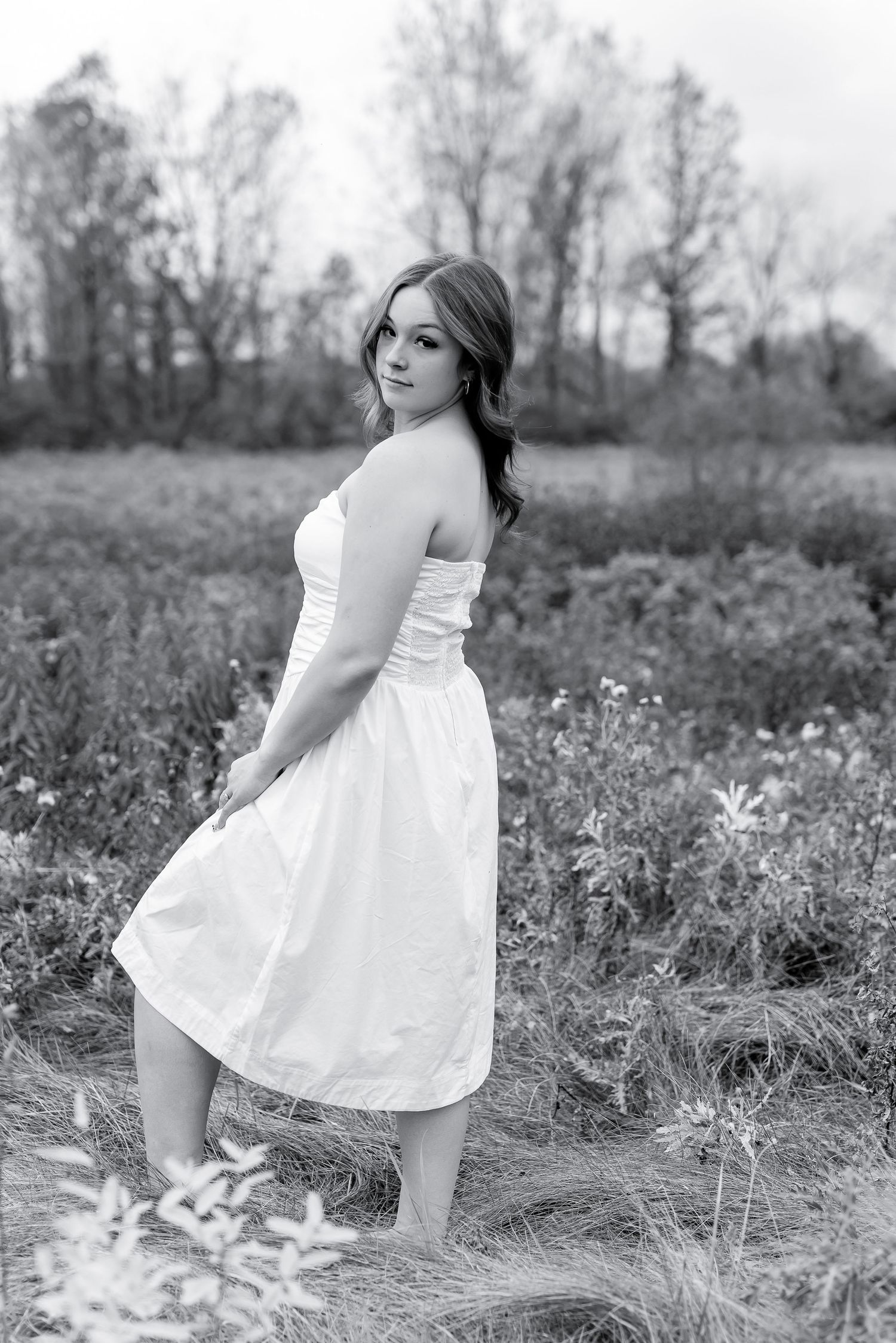 Black and white portrait of young woman in white strapless dress posing in open meadow.
