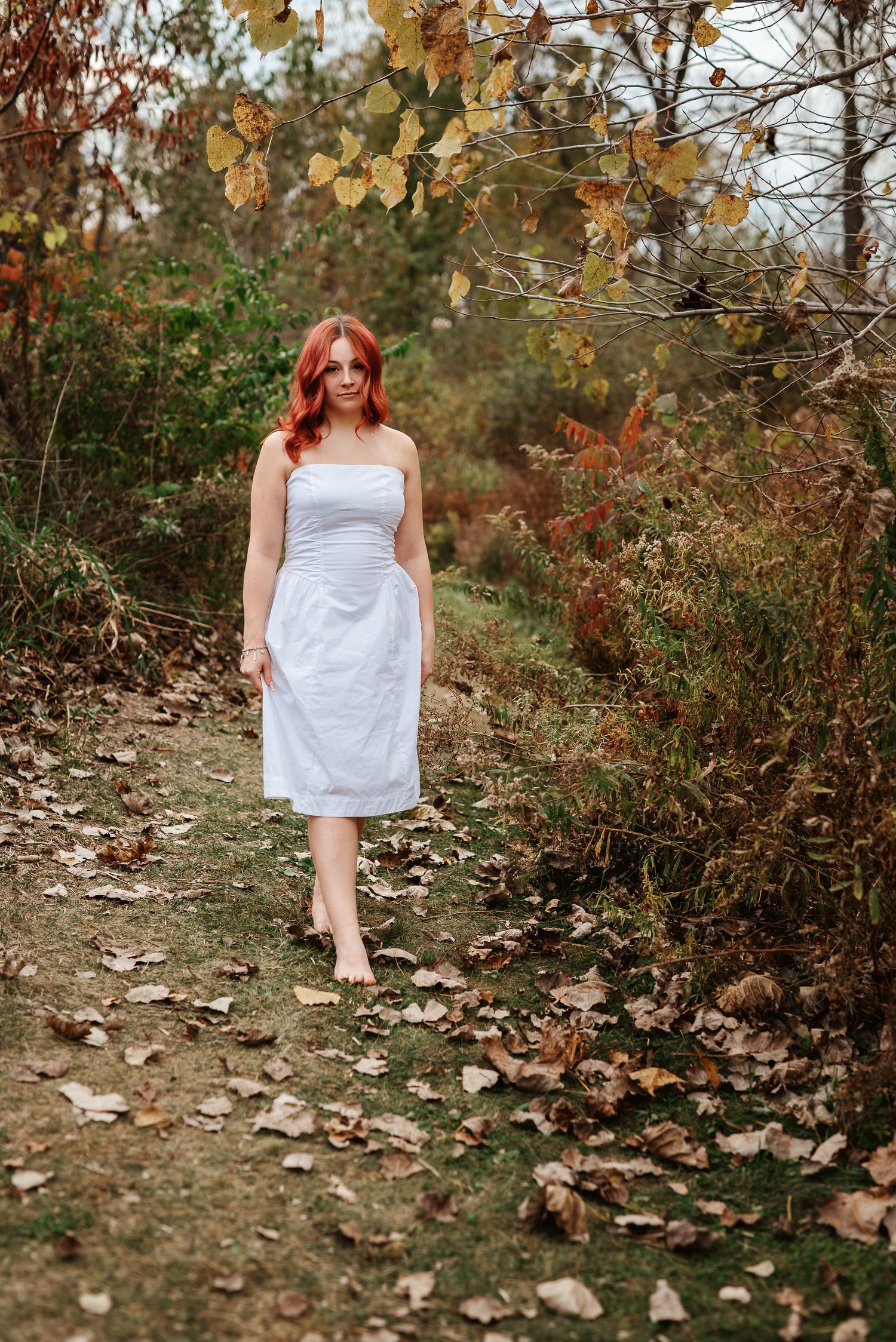 Barefoot redhead woman in white dress walking on leaf-covered path surrounded by fall foliage.