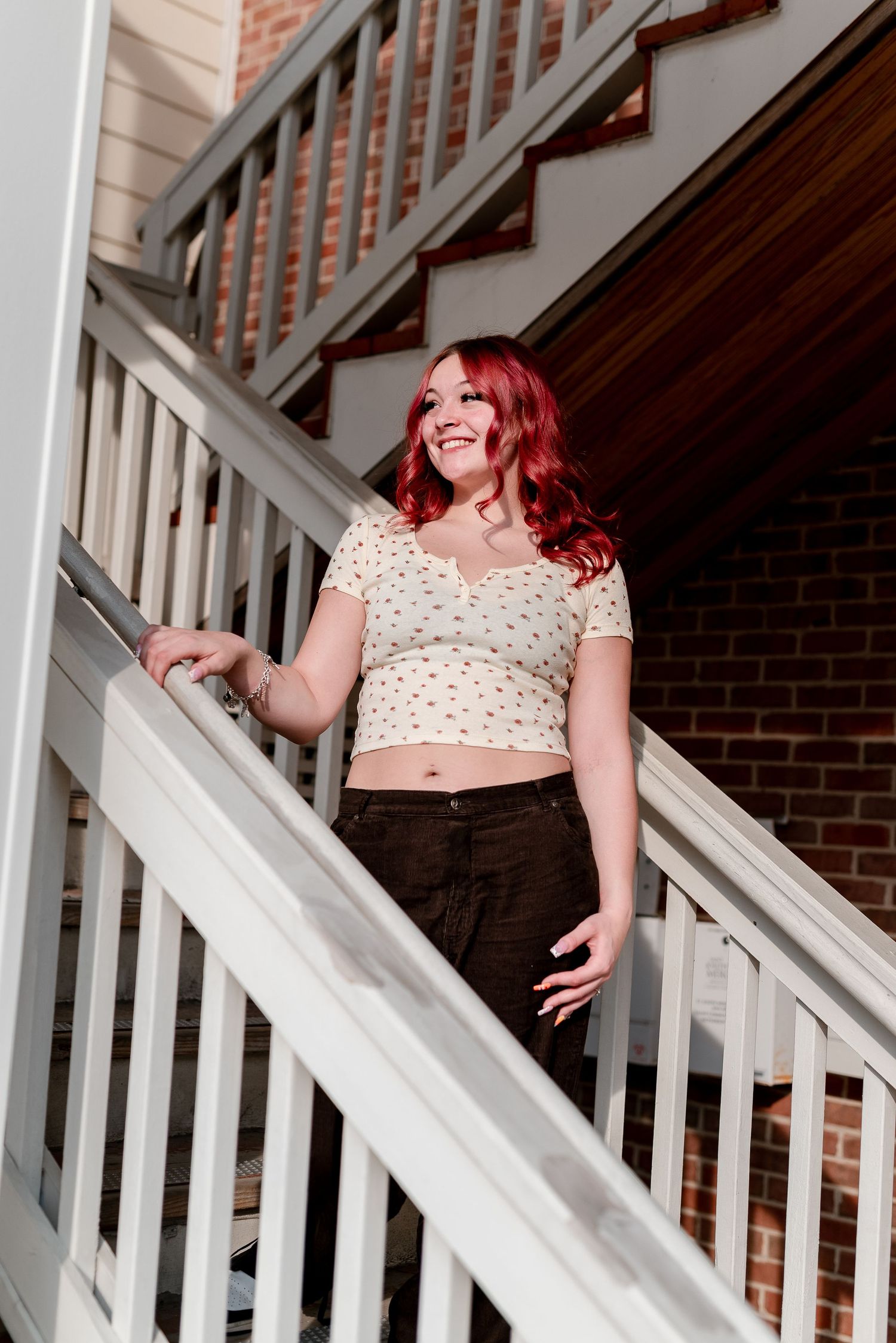 Young woman with red hair smiling on white outdoor staircase wearing crop top and black pants.