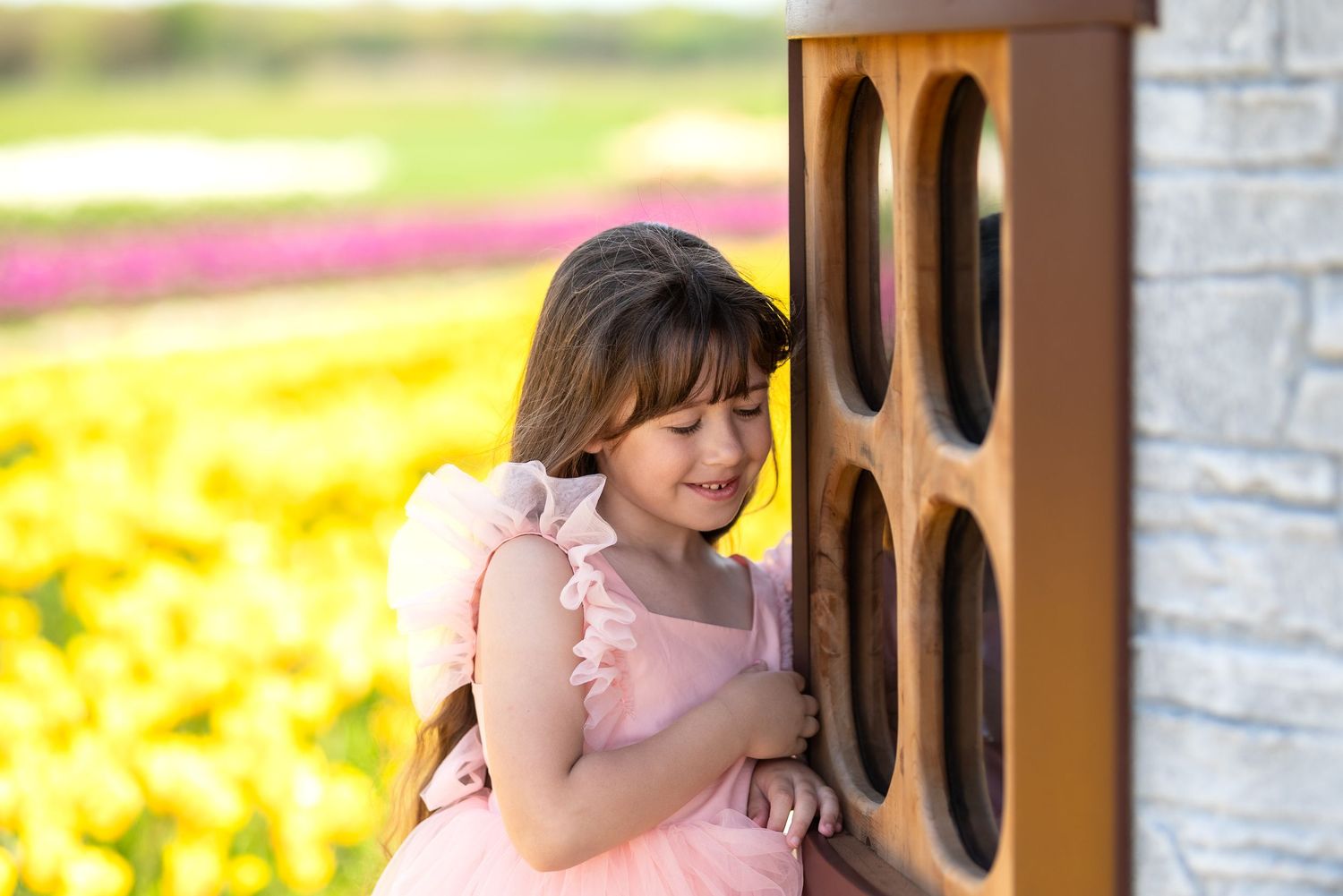 Young girl in pink dress smiling beside wooden structure with colorful tulip fields in background.