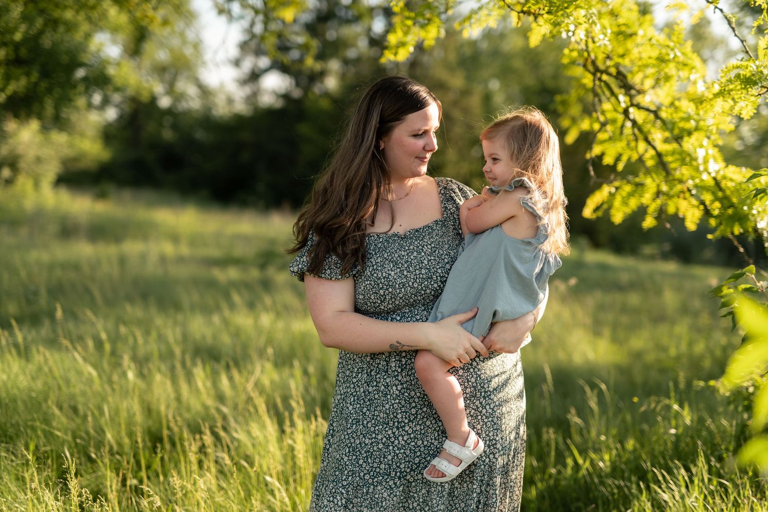 A mother in a floral dress holds her toddler in a sunny green meadow, sharing a joyful moment outdoors.