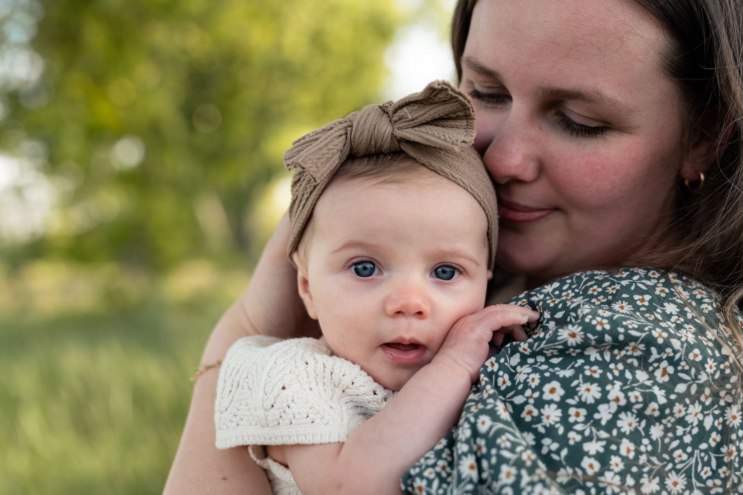A mother tenderly kisses her blue-eyed baby girl wearing a bow headband outdoors in soft natural light.
