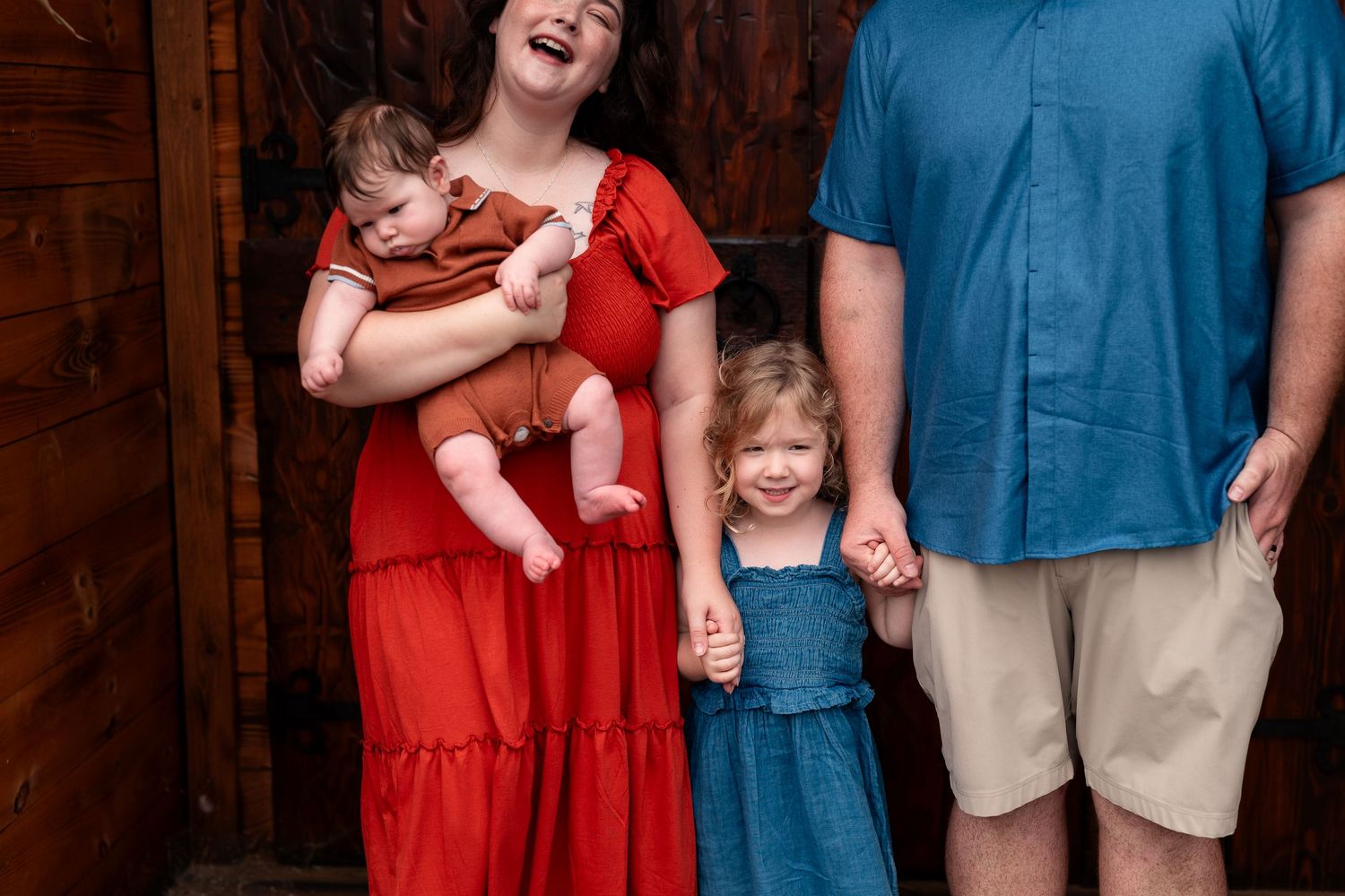 Family of four posing indoors, mother in red dress holding baby, father in blue shirt with toddler girl in denim dress.