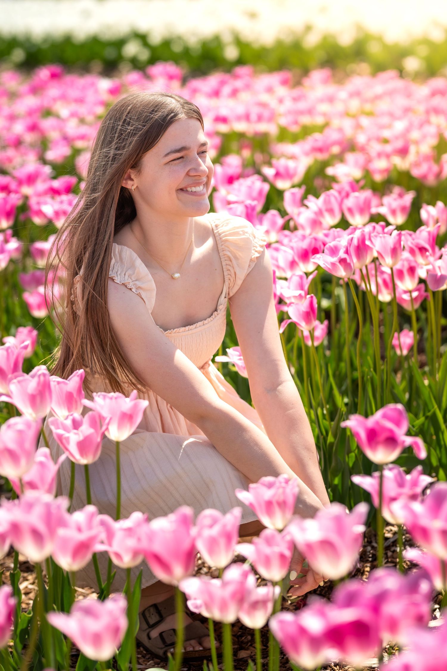 A smiling young woman in a pink dress sits among vibrant pink tulips in a blooming flower field on a sunny day.