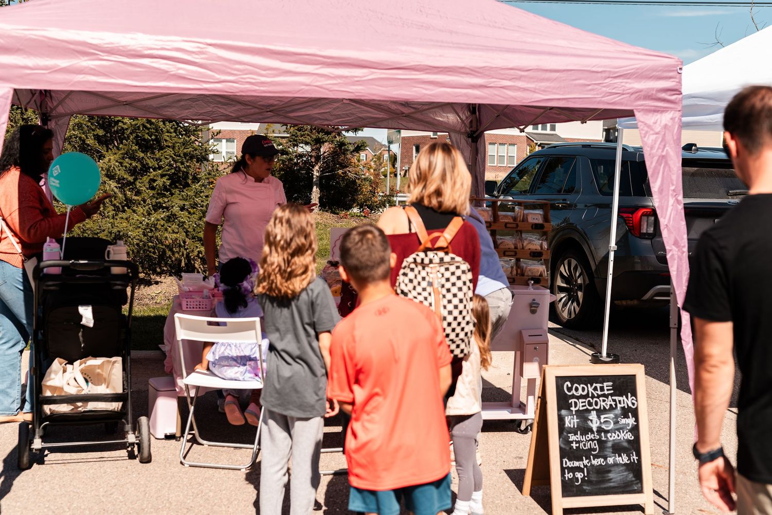 Children and adults shopping at a Cookie Decorations vendor booth under a pink tent at an outdoor fair.