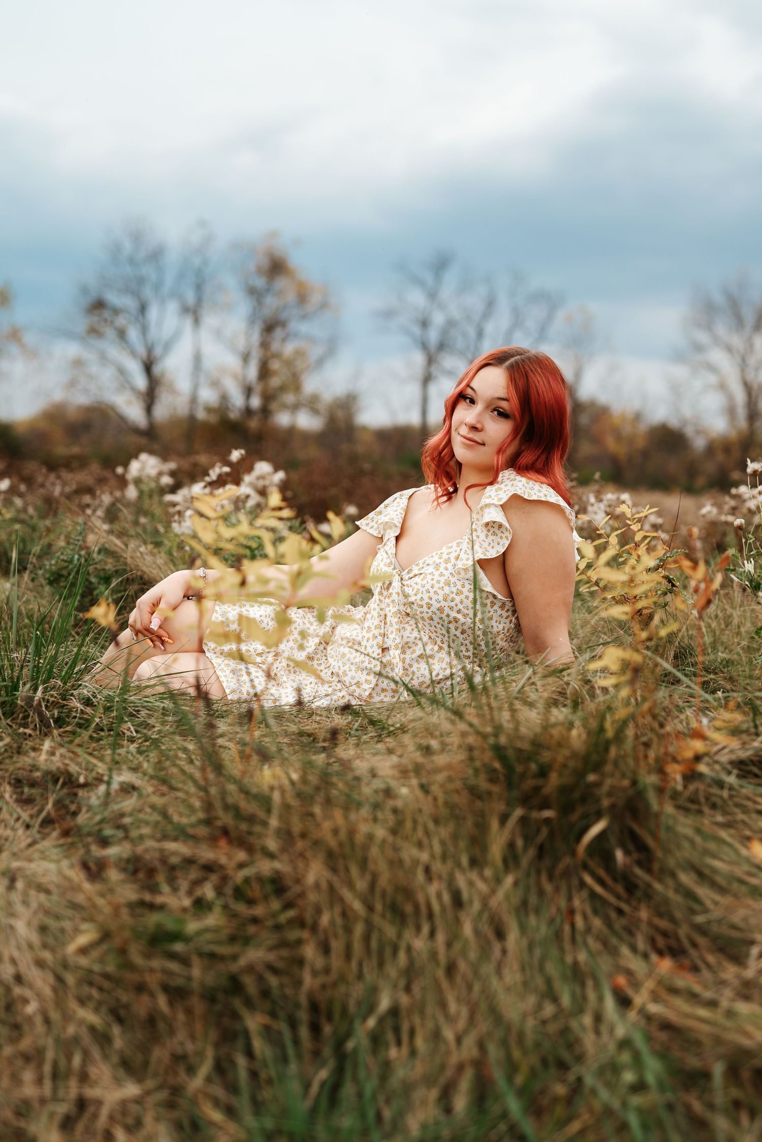 Young woman with red hair in floral dress sitting in autumn field with wildflowers and cloudy sky.