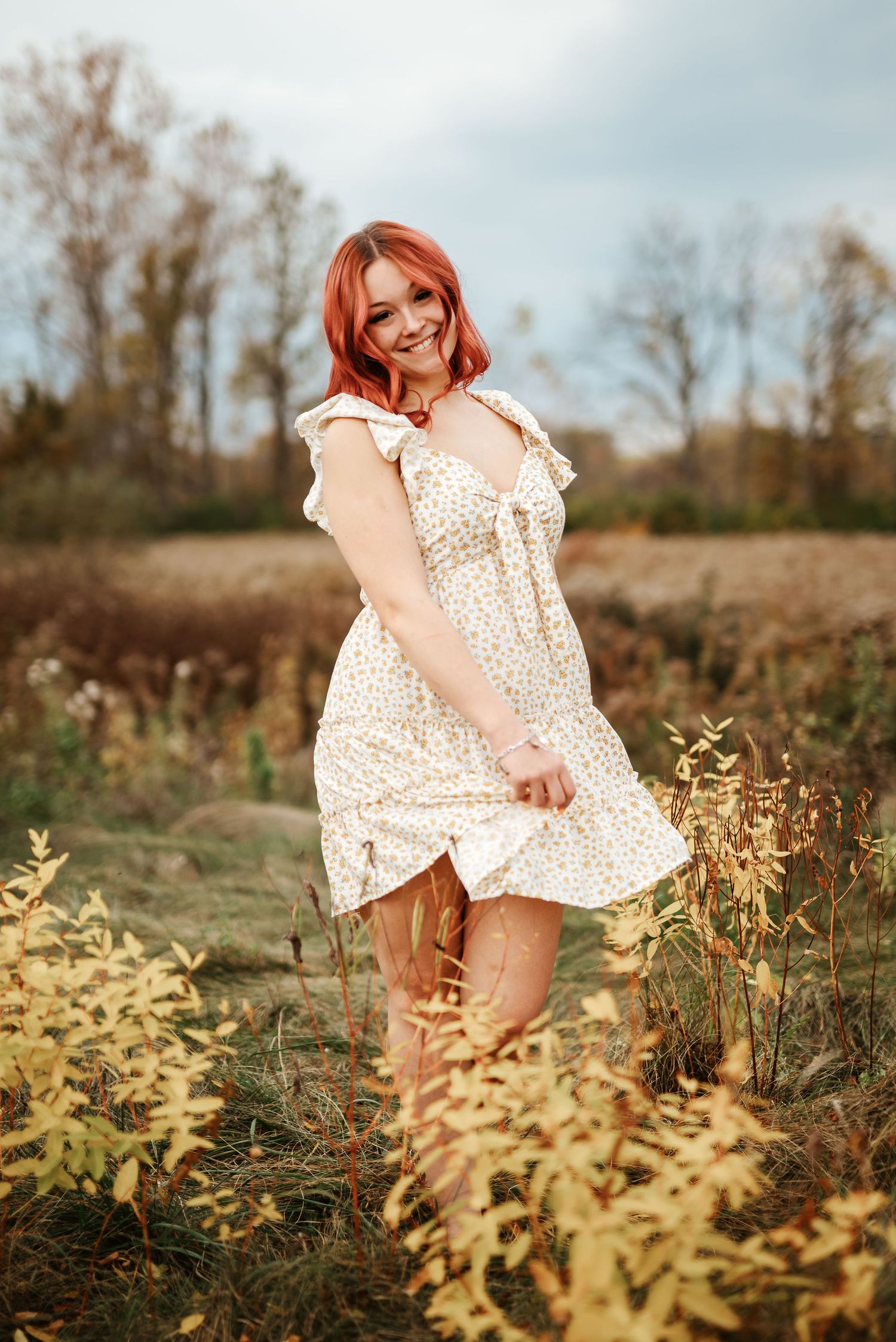 Smiling redhead woman in floral sundress standing in a golden autumn field surrounded by wildflowers.