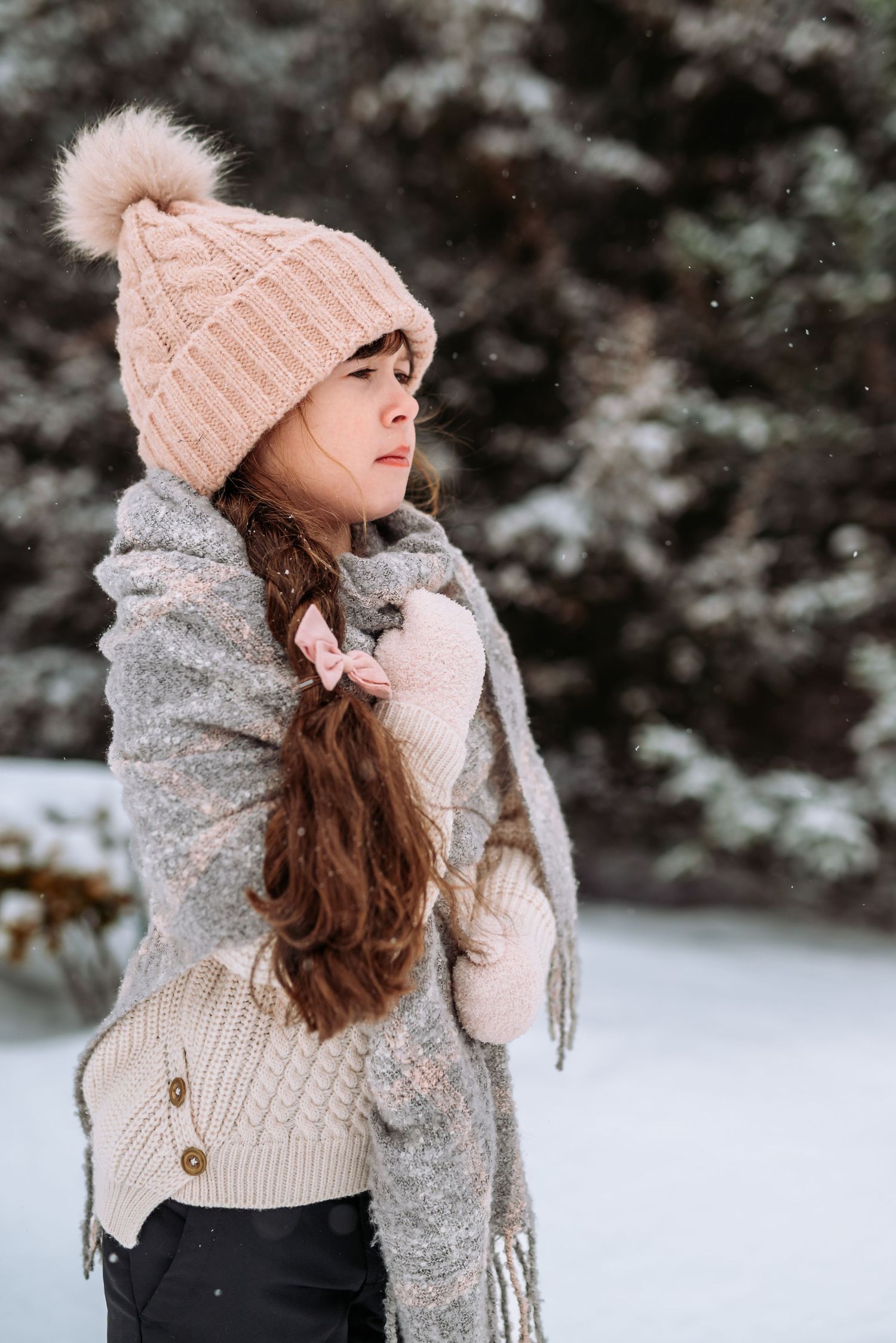 A person in a cozy winter outfit with pink beanie and gray scarf poses against a snowy evergreen background.