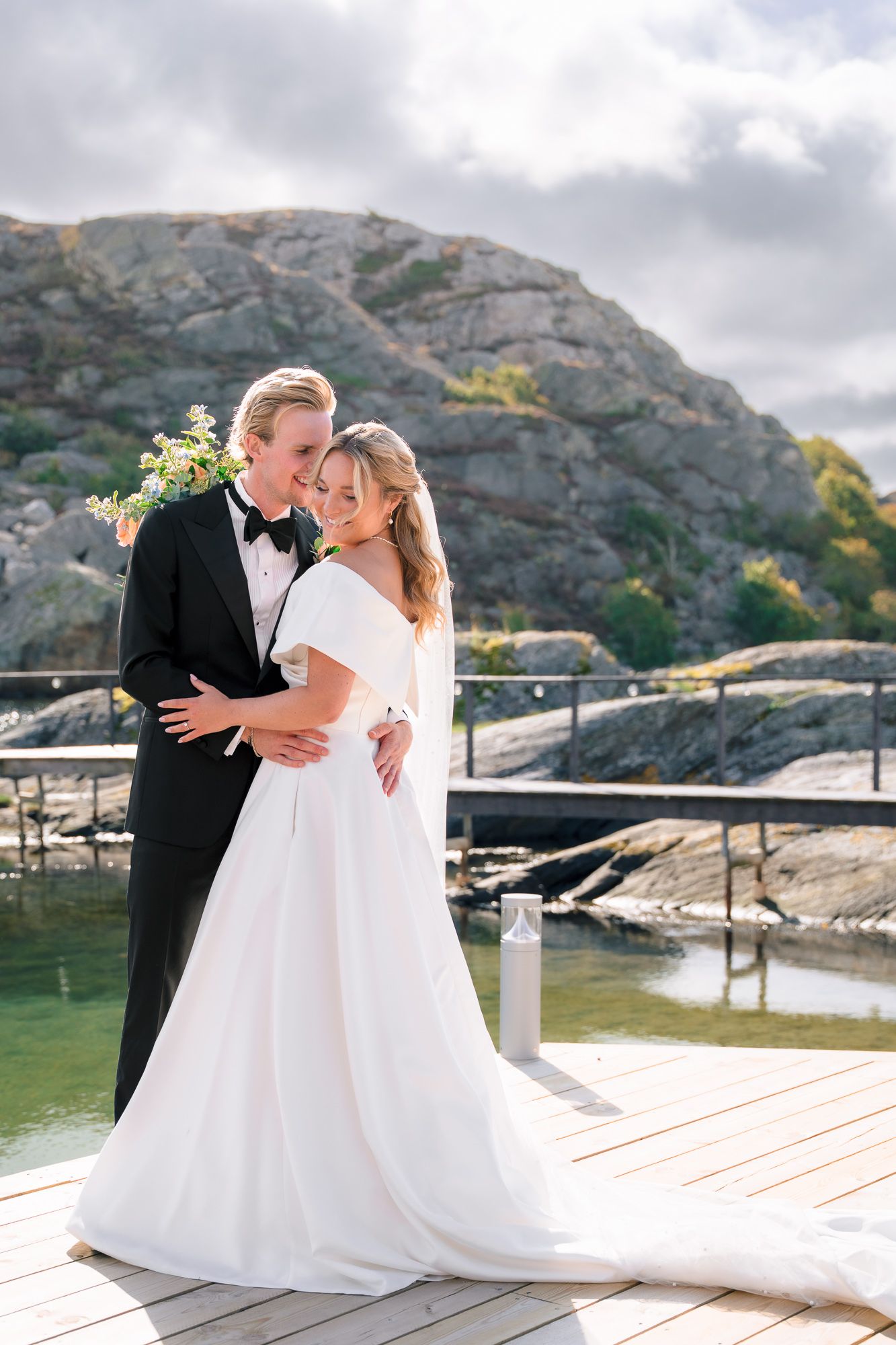 Romantic wedding couple shares intimate moments on a wooden dock with rocky cliffs and water in the background.