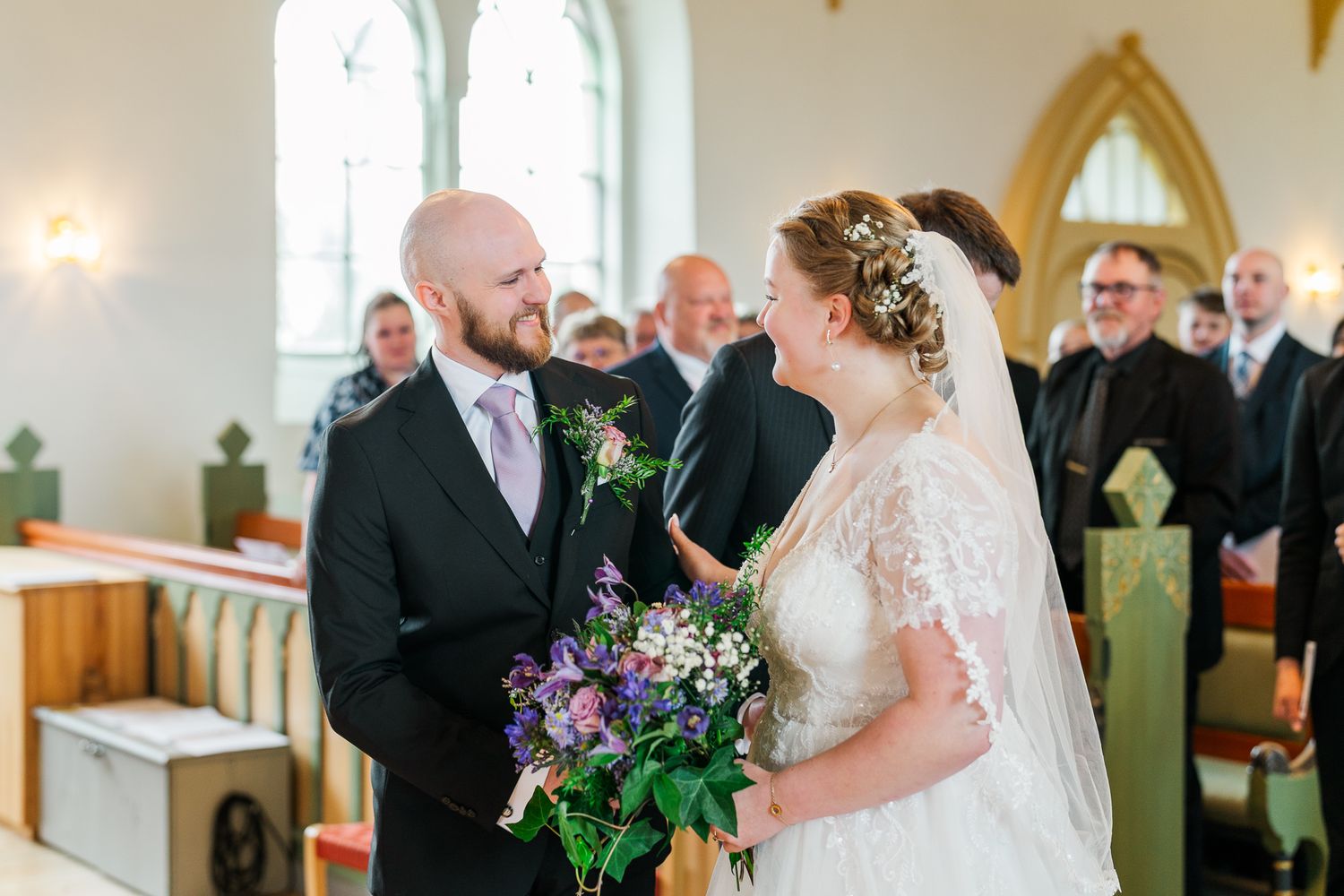 A joyful moment captured between the newlyweds inside a charming church with purple floral bouquet details.