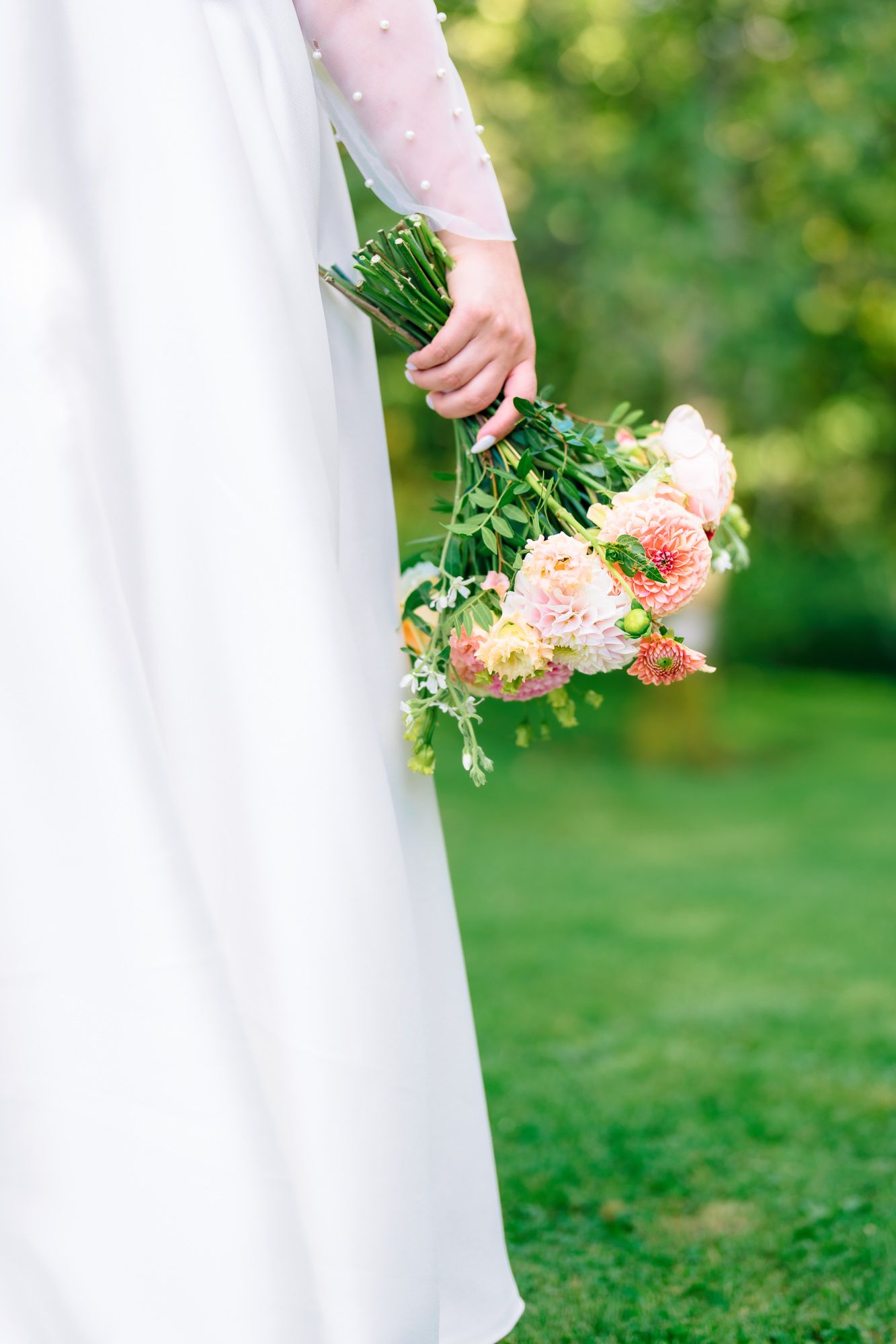 Hand in white dress holds delicate pastel pink bouquet against blurred green garden background.