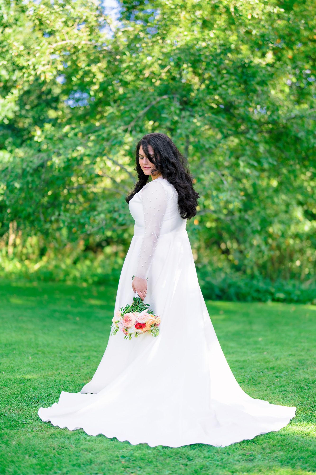 A bride in a flowing white long-sleeve wedding gown holds a floral bouquet while standing on green grass surrounded by trees.