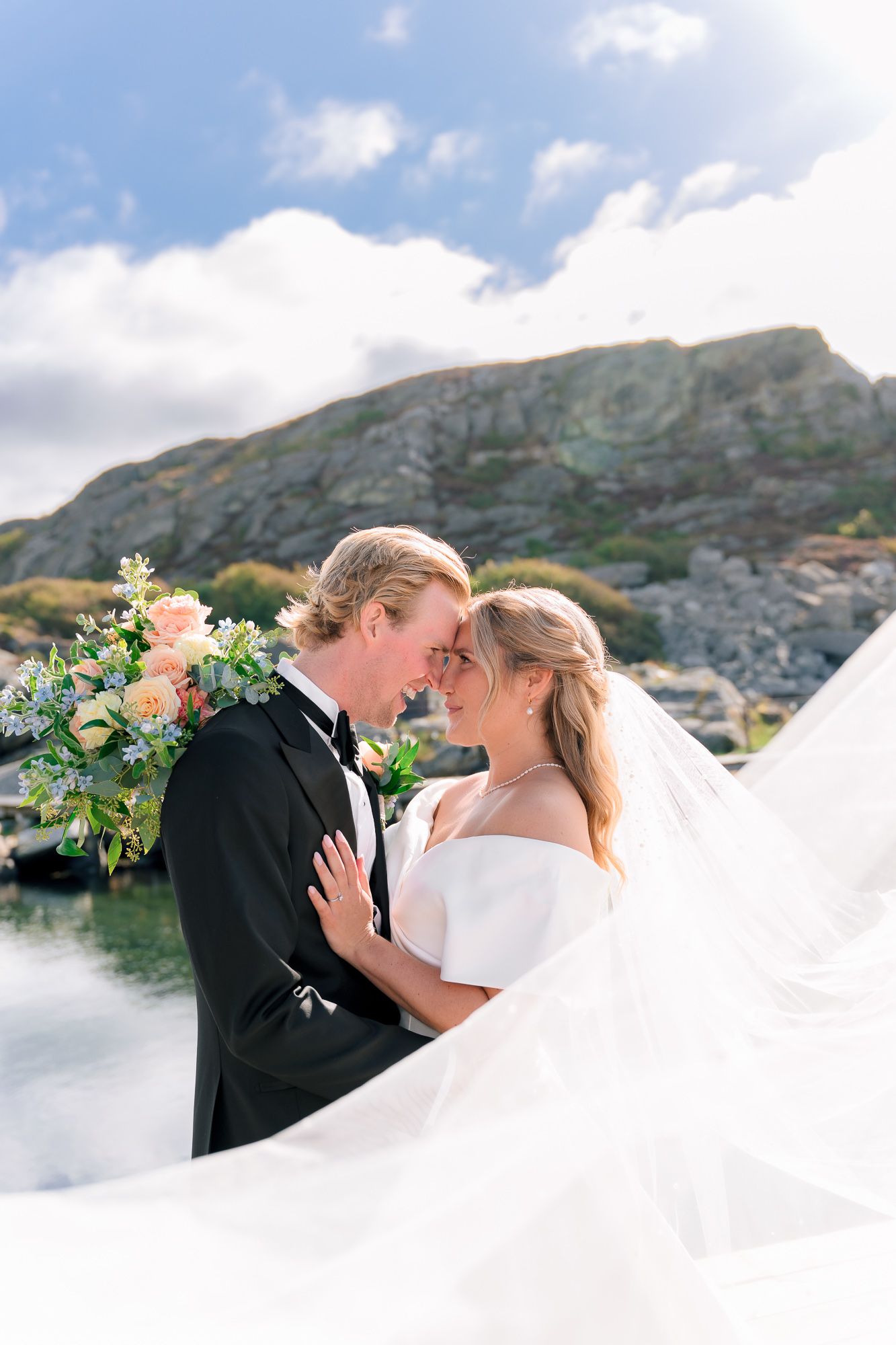 Romantic wedding couple embraces against scenic mountain backdrop with flowing white dress and floral bouquet.