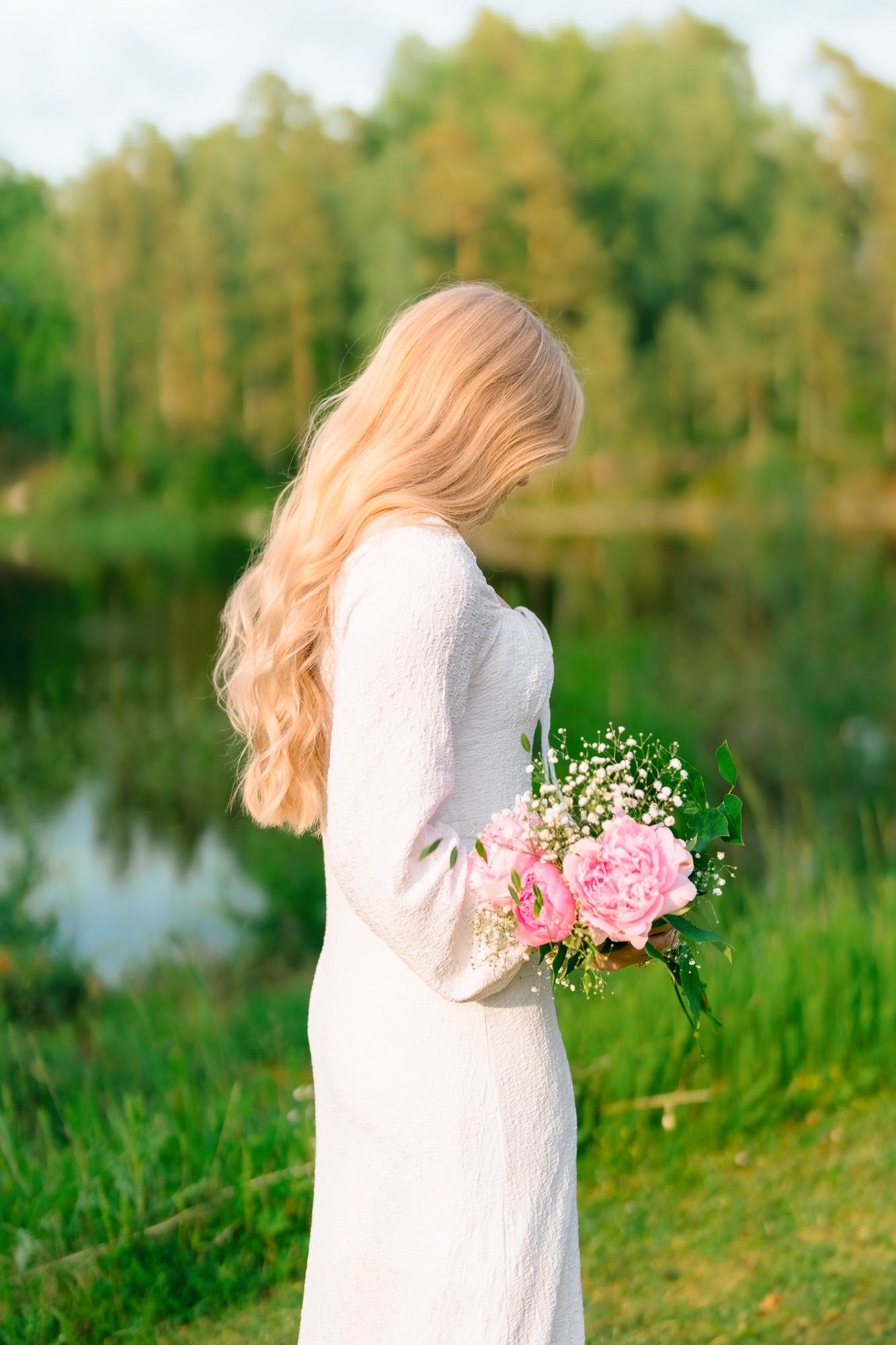 Woman in white dress holding pink peonies and baby's breath flowers stands by a tranquil lake at sunset.
