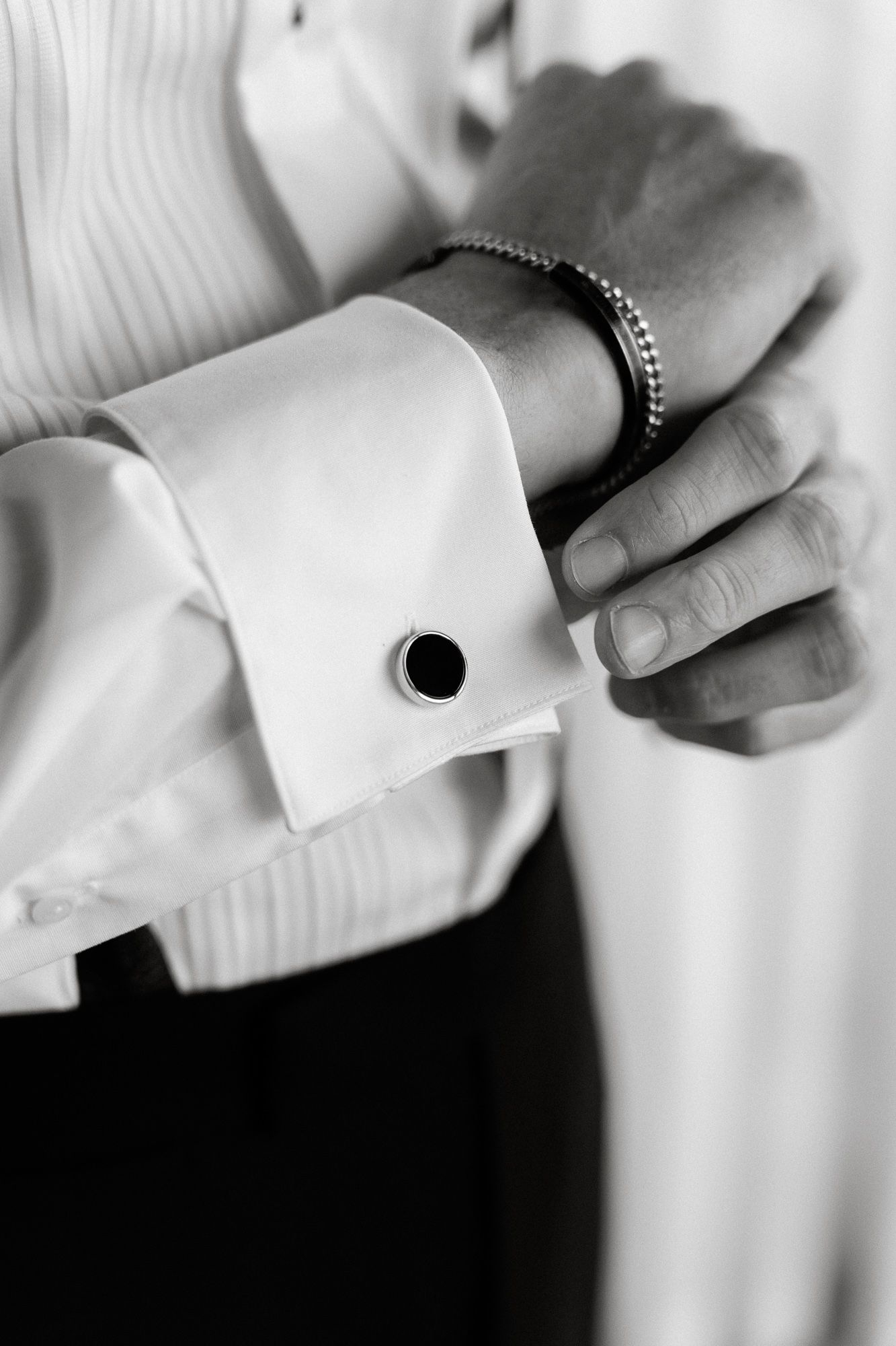 A black and white close-up of a formal white shirt cuff with cufflink and bracelet on wrist.