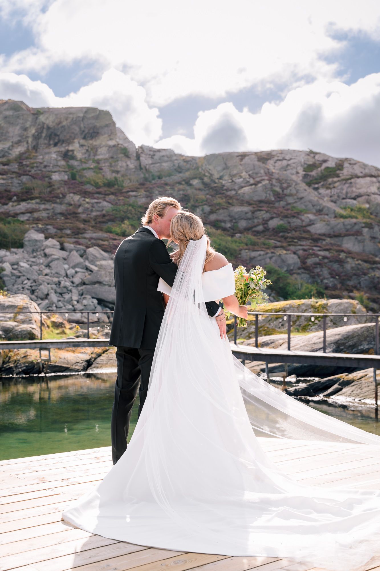 A couple shares an intimate moment on a wooden dock overlooking scenic rocky cliffs and calm waters.