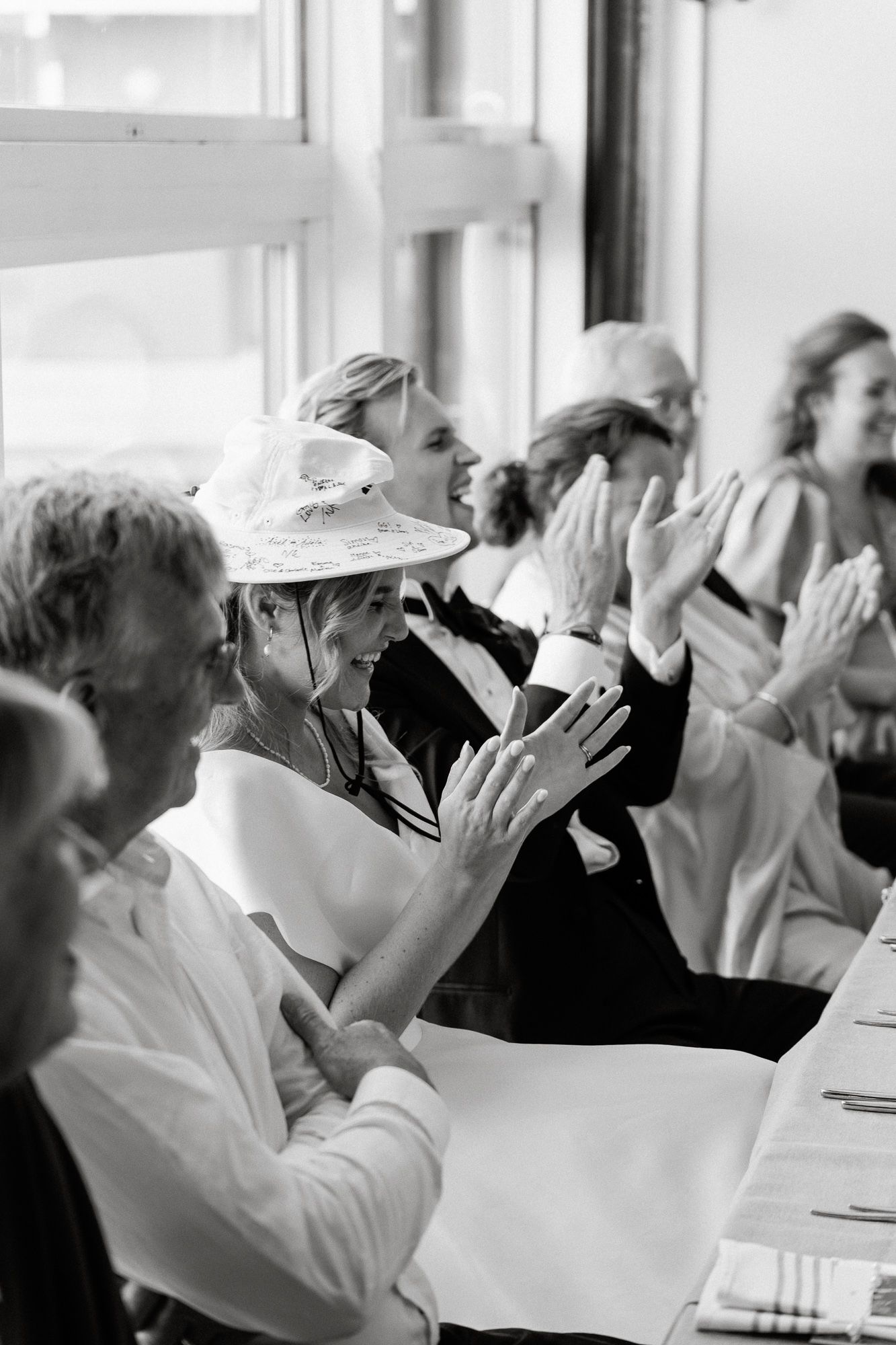 A black and white image shows a row of people in white uniforms sitting at a table and raising their hands in unison.