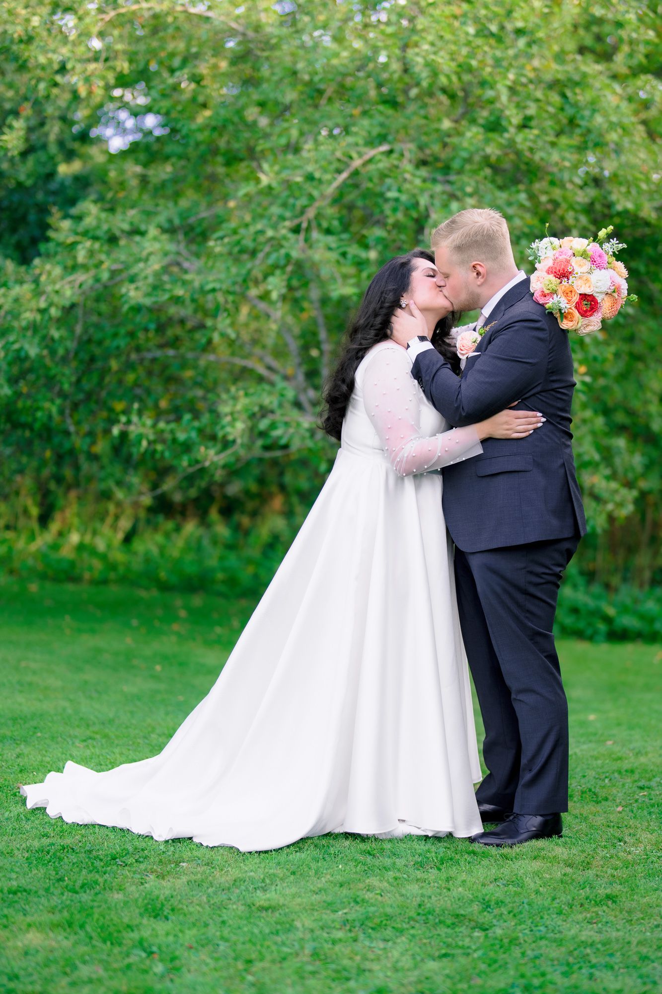 Couple shares romantic kiss on green lawn while bride holds vibrant pink and coral bouquet against lush garden backdrop.