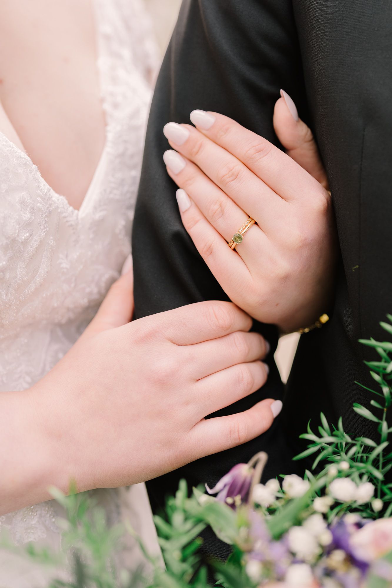 Close-up of hands with wedding ring against black suit and white lace dress with purple flowers in background.