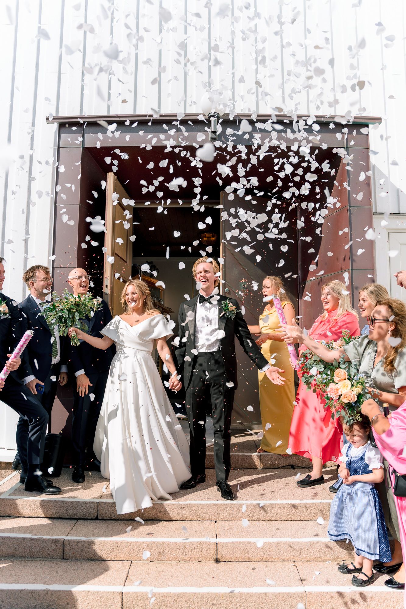A wedding party celebrates on church steps as white confetti rains down during a joyous exit celebration.