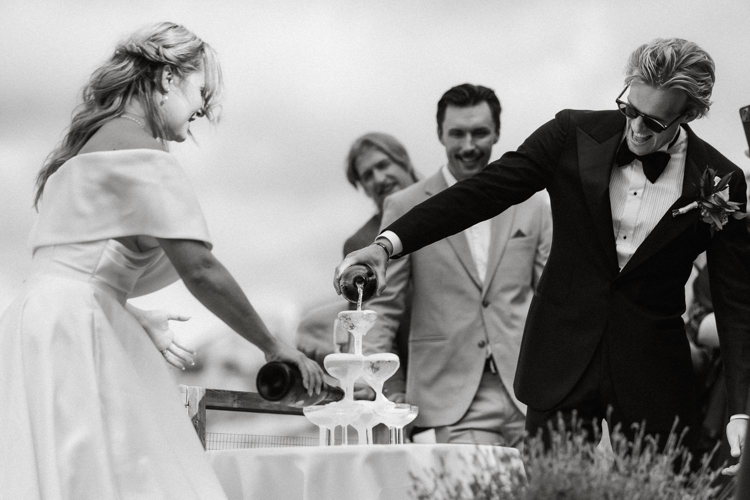 Wedding guests gather around a champagne tower while pouring bubbly drinks during an elegant celebration.