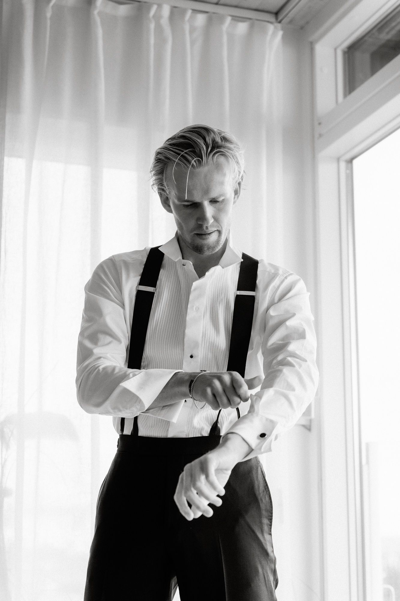 A person in formal attire with black suspenders and white shirt adjusts their sleeves by a bright window.