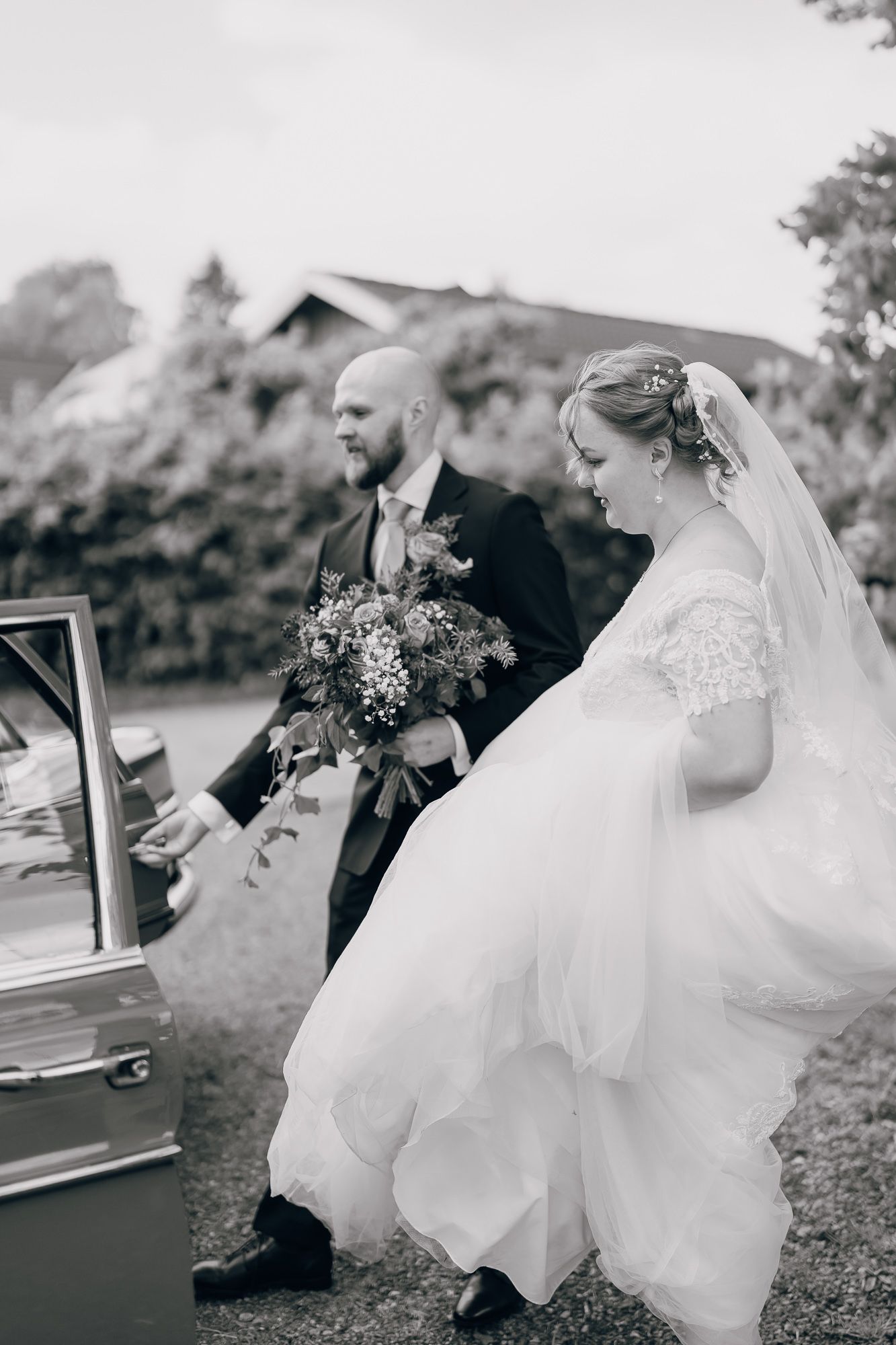 Black and white wedding photo showing couple getting into vintage car with bride wearing lace gown and veil.