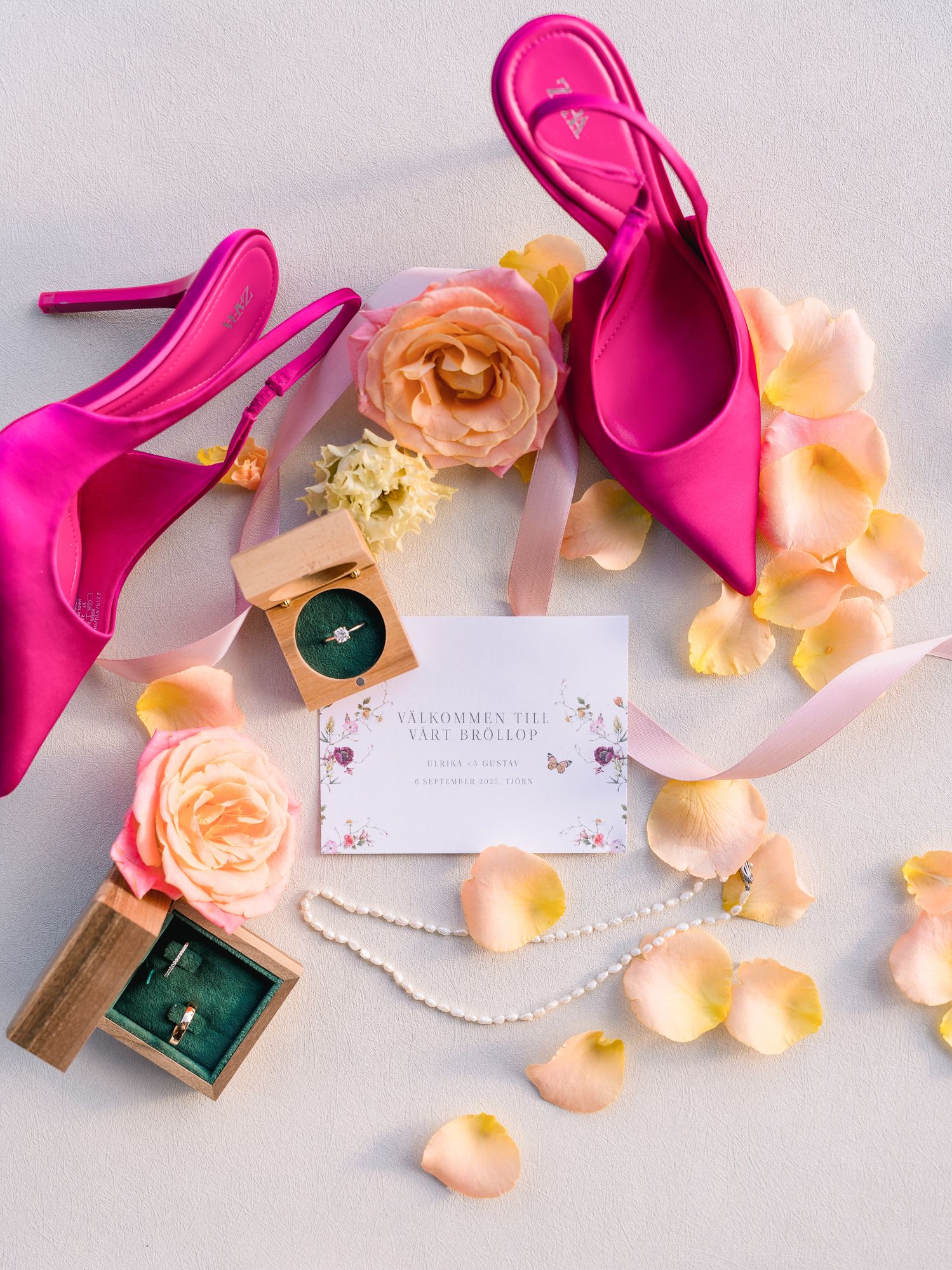 Pink high heel shoes and peach rose petals arranged with pearl necklace and emerald jewelry box on white surface.