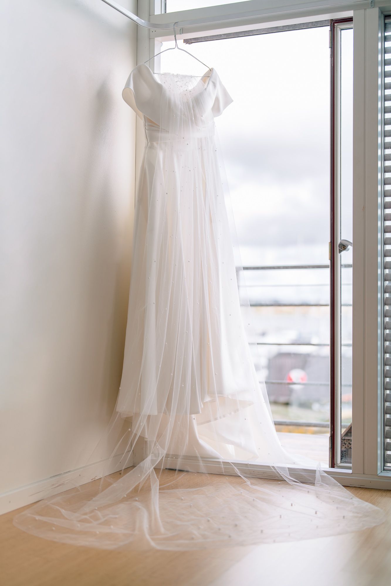 White wedding dress hanging by window with natural light streaming through sheer fabric.