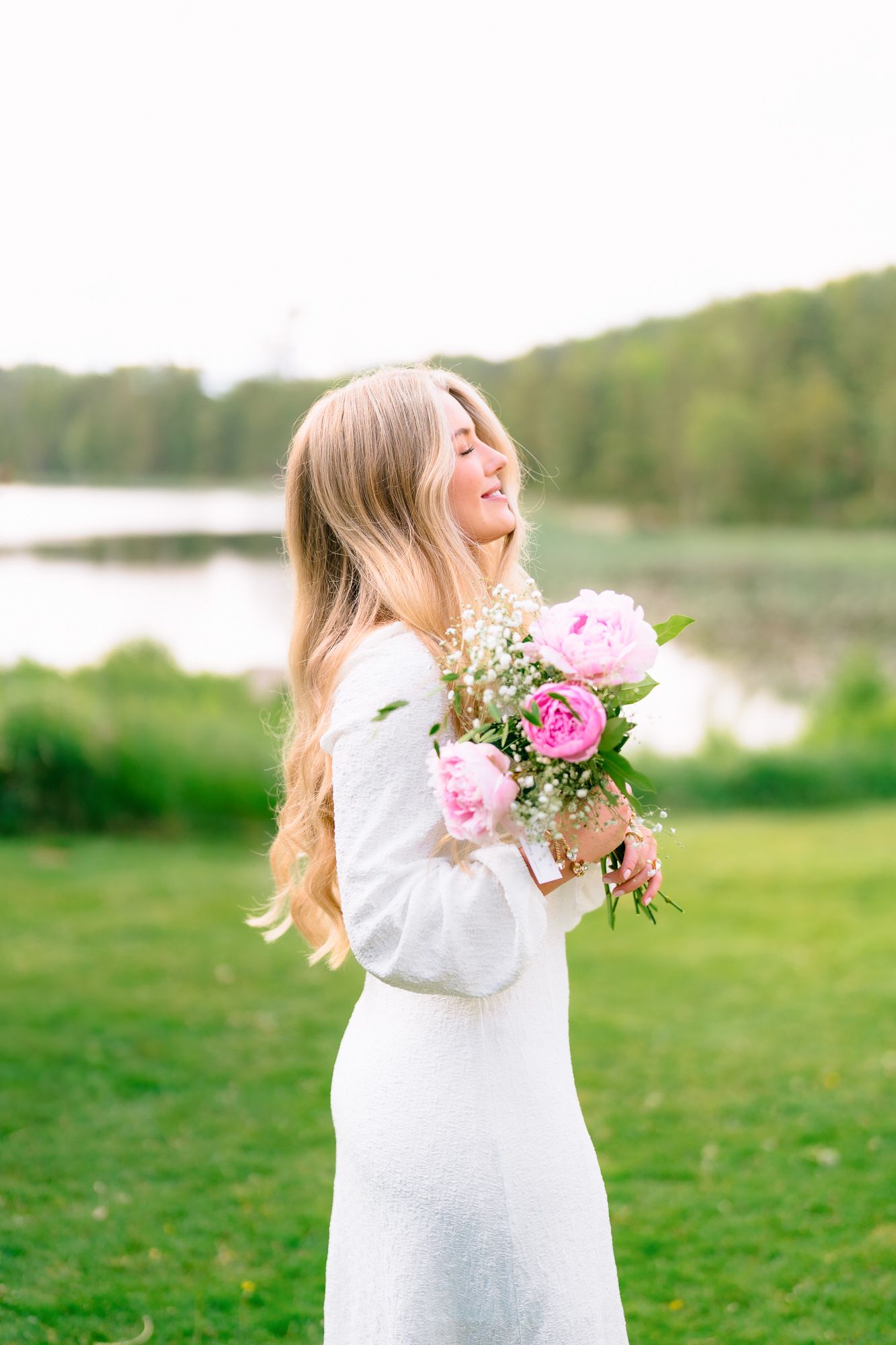 Woman in white dress holds pink peony bouquet while standing near lake and smiling on sunny day.