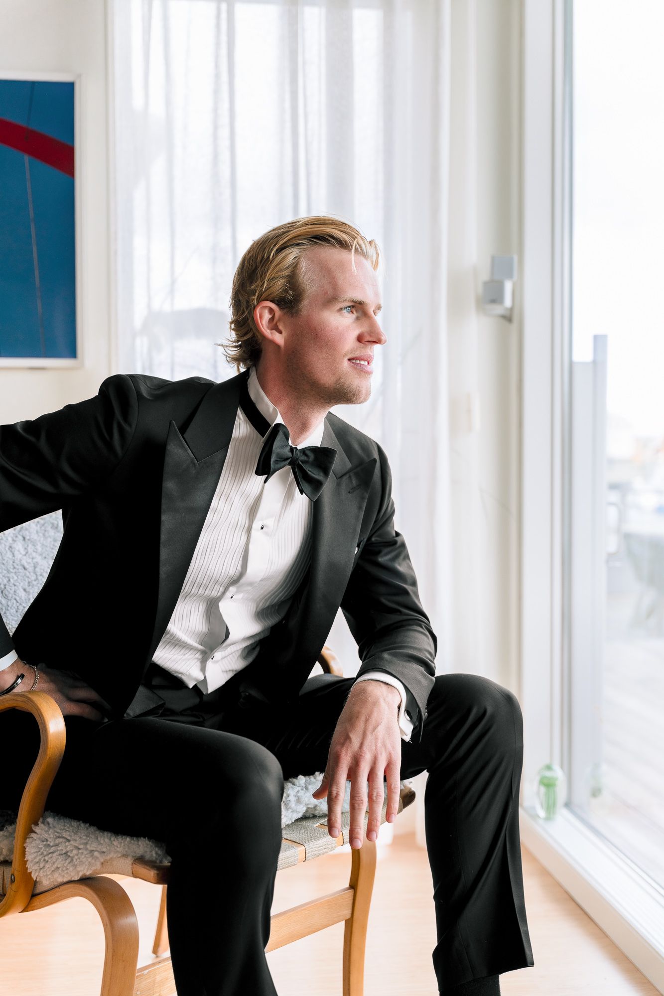 A person in a black tuxedo and white shirt sits thoughtfully on a wooden chair by large windows.